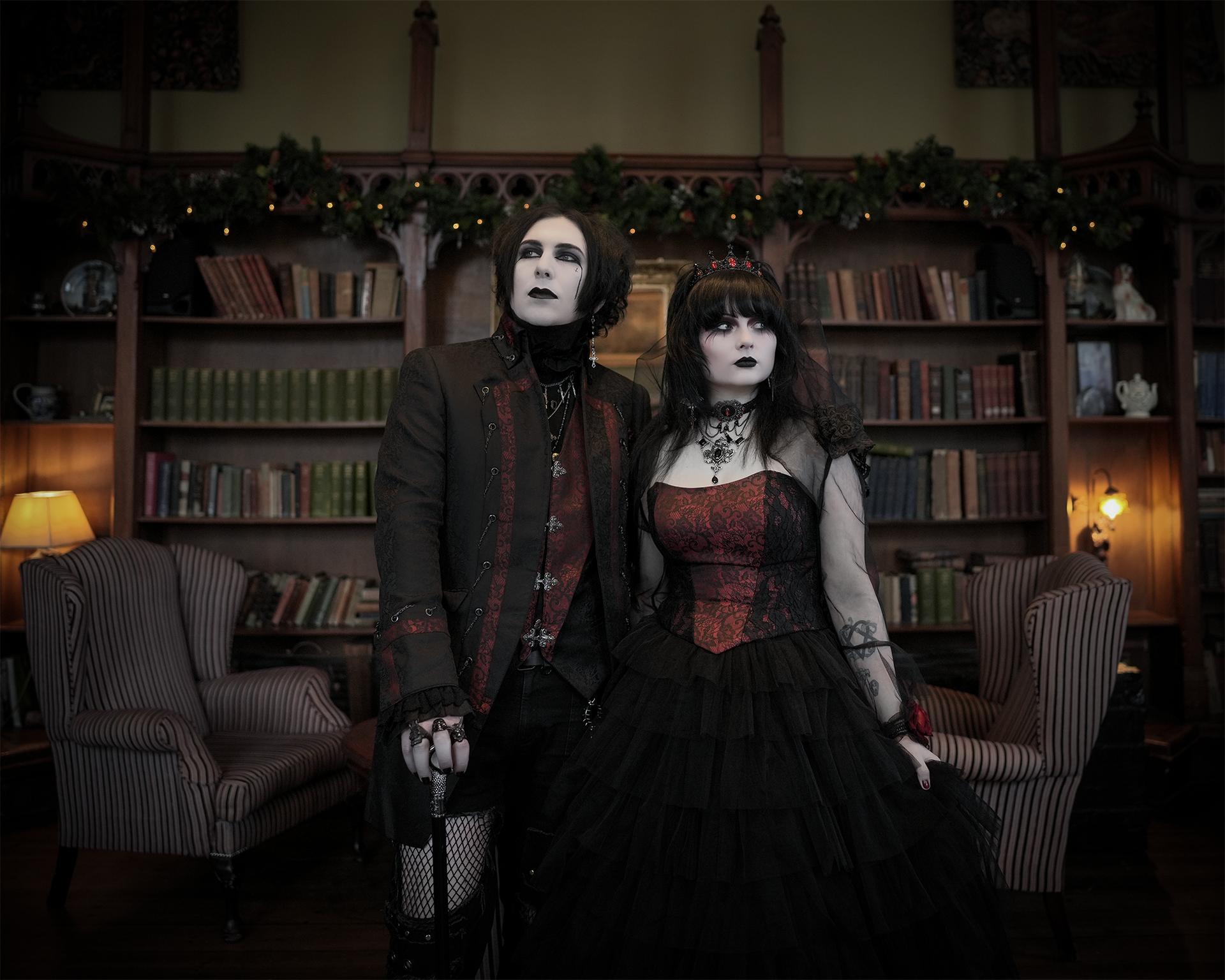 Ein Paar steht in einer urischen Bibliothek. Die Frau trägt ein aufwendig gestaltetes, schulterfreies Brautkleid im Gothic-Stil. Das Oberteil und ein Teil des Rockes sind in dunklem Rot mit schwarzem Brokatmuster gehalten, der restliche Rock ist schwarz und voluminös. Sie hält ihren Rock mit einer Hand fest, ein hauchdünner Bolero aus schwarzem Tüll bedeckt ihre Schultern und Arme. Ihr langes, dunkles Haar ist mit Ponny, teils offen und fällt auf die Schulter. Sie trägt auffälliges, dunkles und blasses Make-up mit schwarzen Akzenten sowie ein schwarzes Halsband mit einem roten Stein und auf dem Kopf eine Krone in schwarz mit roten Steinchen. Der Mann steht rechts neben ihr, trägt einen schwarzen Gehrock mit roten Brokatverzierungen an den Revers und Ärmelabschlüssen und Ketten. Die Verzierungen sind passend zum Kleid in dunkel rot, die Weste hat ebenfalls schwarz-rote Akzente und elegante Verschlüsse. In der Hand hält er einen Gehstock und hat auffallend freizügige Hosen mit Netz und nackter haut. Alles in dunkel und schwarz gehalten. Er hat dunkles, voluminöses Haar bis über die Ohren und ohne Bart. Beide halten sich gedrückt an einander und schauen jeweils links und rechts und nicht in die Kamera. Die Stimmung wirkt romantisch, stilvoll und leicht mystisch.
Dieses Outfit ist im Gothic-Style gehalten und erhältlich im Dark Dream Atelier für Gothic- und Steampunk-Mode in Gründau.