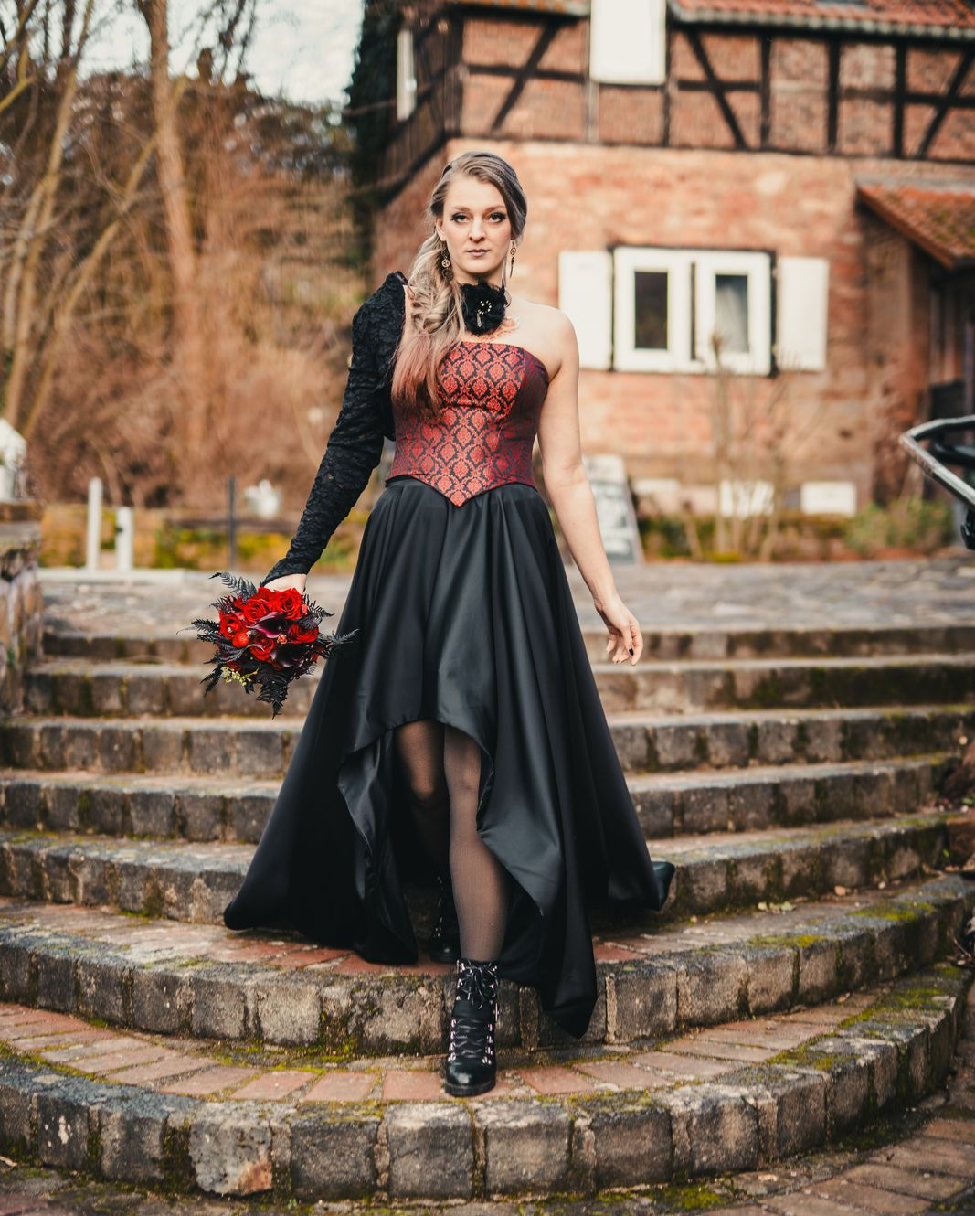 Frau in einem schwarz-roten Gothic-Kleid mit asymmetrischem Rock, Korsage mit Brokatmuster und einer schwarzen Spitzenmanschette am rechten Arm. Sie hält einen Strauß roter Rosen und steht auf steinernen Stufen vor einem Fachwerkhaus.
Die gezeigten Fantasy- und LARP-Outfits sind erhältlich bei Mr. & Mrs. Huebner – Dark Dream Atelier in Gründau