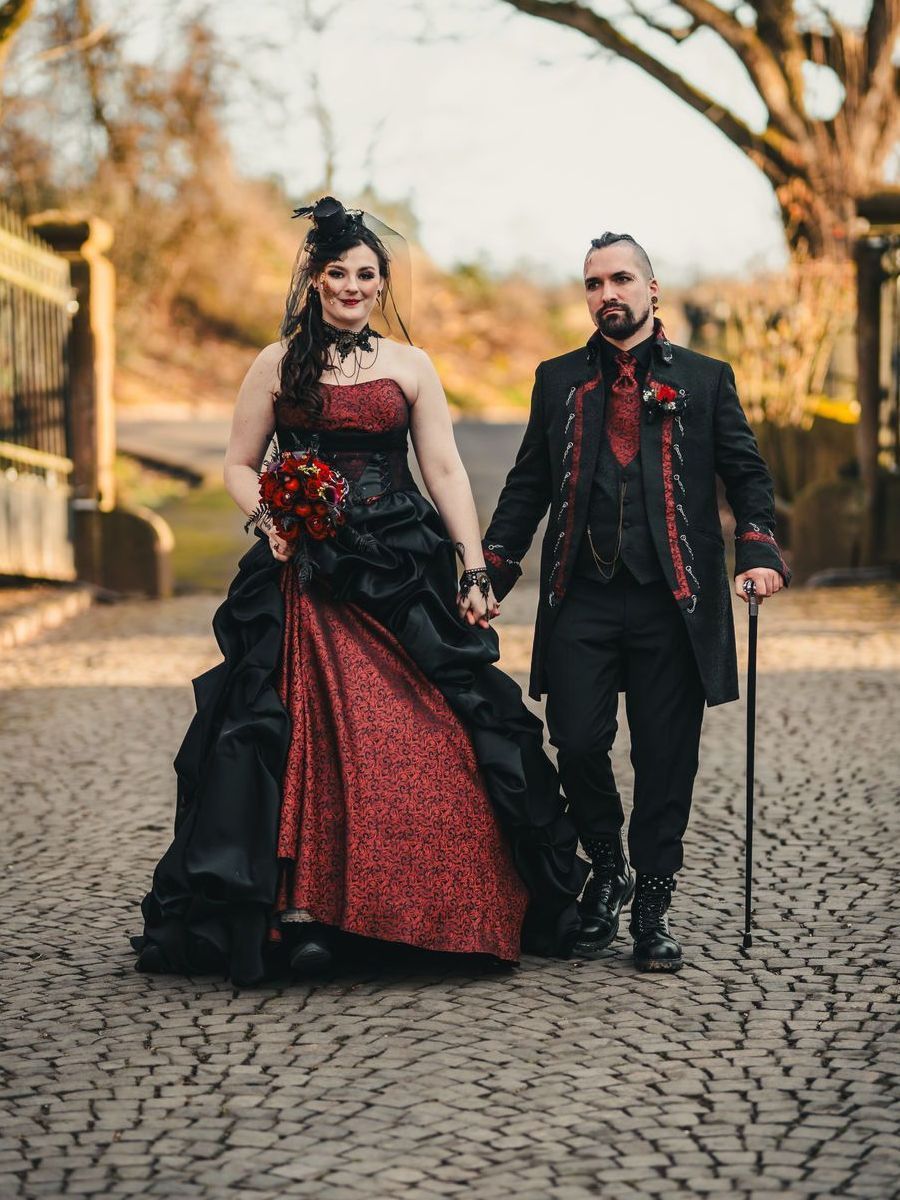 Das Bild zeigt ein Hochzeitspaar in aufwendig gestalteter, alternativer Festkleidung im Stil der Steampunk Art, wie sie für ausgefallene Hochzeiten oder Fantasy-Events typisch ist. Die Szene spielt im Freien auf einem historischen, gepflasterten Weg zwischen alten Torpfosten mit Blick auf eine herbstlich kahle Landschaft.
Links im Bild geht eine Frau mit aufwendig gestyltem, dunklem Haar, das seitlich gesteckt ist und von einem kleinen schwarzen Fascinator mit Schleier geziert wird. Sie trägt ein barock inspiriertes, schwarzes und bordeauxrotes Brautkleid mit Korsage und üppigem Rock. Die Korsage ist ärmellos und aus bordeauxfarbenem Stoff mit schwarzem Muster gearbeitet. Der Rock besteht aus mehreren Lagen mit schwarzen Drapierungen über einem roten, gemusterten Unterrock. Sie hält einen passenden Brautstrauß aus roten und schwarzen Rosen mit dunklem Blattwerk.
Rechts neben ihr geht ein Mann mit Bart, Undercut-Frisur und auffälligen Ohrringen. Er trägt ein dreiteiliges, schwarzes Ensemble mit rot-schwarzen Steampunk-Details: Eine Brokatweste, ein Stehkragenhemd mit rot abgesetzten Knopfleisten, ein langes Gehrock-Jackett mit Schnürungen und dekorativen Nähten. An seiner rechten Hand hält er einen schwarzen Spazierstock. Seine Schuhe und Accessoires wirken punkig und individuell.
Der Ausdruck beider ist stolz, das Bild vermittelt Selbstbewusstsein, Individualität und Liebe zum Detail. Der Look beider ist ein Beispiel für moderne Steampunk Art Couture, bei der viktorianische Elemente mit düsterer Eleganz und moderner Interpretation verschmelzen.
Die gezeigten Outfits stammen aus der Steampunk-Art-Kollektion von Mr. & Mrs. Hübner – Dark Dream Atelier in Gründau