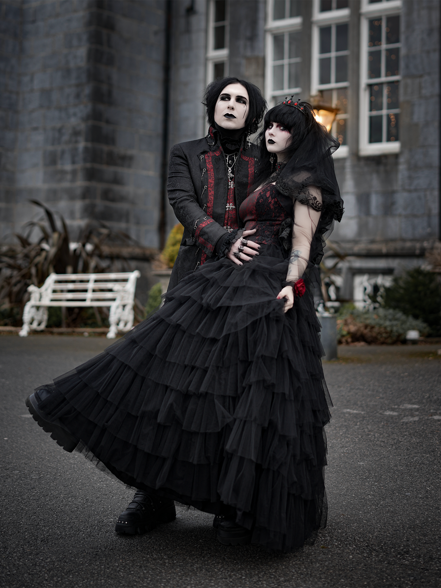 Das Bild zeigt ein Paar in expressiver, schwarzer Gothic-Hochzeitsmode vor einem historischen Gebäude mit grau-blauer Steinfassade und weißen Fenstern. Sie stehen auf einer nassen Asphaltfläche, vermutlich nach einem Regenschauer. Im Hintergrund ist eine weiße Bank und etwas herbstlich verwelktes Grün zu sehen. Die Lichtstimmung ist dramatisch und unterstreicht den düsteren, romantischen Stil der Szene.
Der Mann auf der linken Seite trägt ein langes schwarzes Jackett mit roten Brokatdetails an Kragen, Revers und Manschetten. Unter dem Jackett trägt er eine schwarze Bluse mit Halsband und Schmuckdetails. Sein Gesicht ist blass geschminkt, die Augenpartie betont. Schwarzes Haar und schwarze Fingernägel ergänzen seinen alternativen Look im Dark Romantic Style.
Die Frau an seiner Seite trägt ein tiefschwarzes, voluminöses Tüllkleid mit mehreren Stofflagen und Rüschen. Das Oberteil besteht aus rotem Brokat mit schwarzer Spitze, kombiniert mit einem schwarzen Schleier, der über Haar und Gesicht fällt. Ihre Arme sind mit Tattoos bedeckt, sie trägt schwarze Lippen und dramatisches Augen-Make-up. Ihr Blick ist melancholisch und ausdrucksstark – der Look erinnert an viktorianische Trauermode, interpretiert im modernen Gothic-Stil.
Das Paar hält sich eng umschlungen und wirkt emotional verbunden, romantisch, ausdrucksstark und individuell.
Die gezeigten Gothic-Hochzeitsoutfits sind erhältlich bei Mr. & Mrs. Hübner – Dark Dream Atelier in Gründau für Berlin