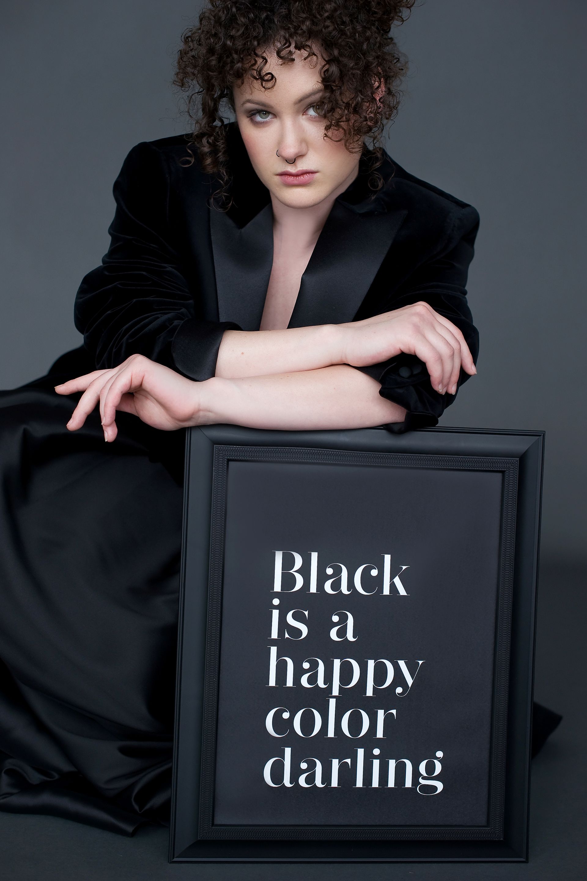 unge Frau mit lockigem Haar und schwarzem Outfit posiert vor dunklem Hintergrund. Vor ihr steht ein schwarzer Bilderrahmen mit weißer Schrift: ‚Black is a happy color darling‘.