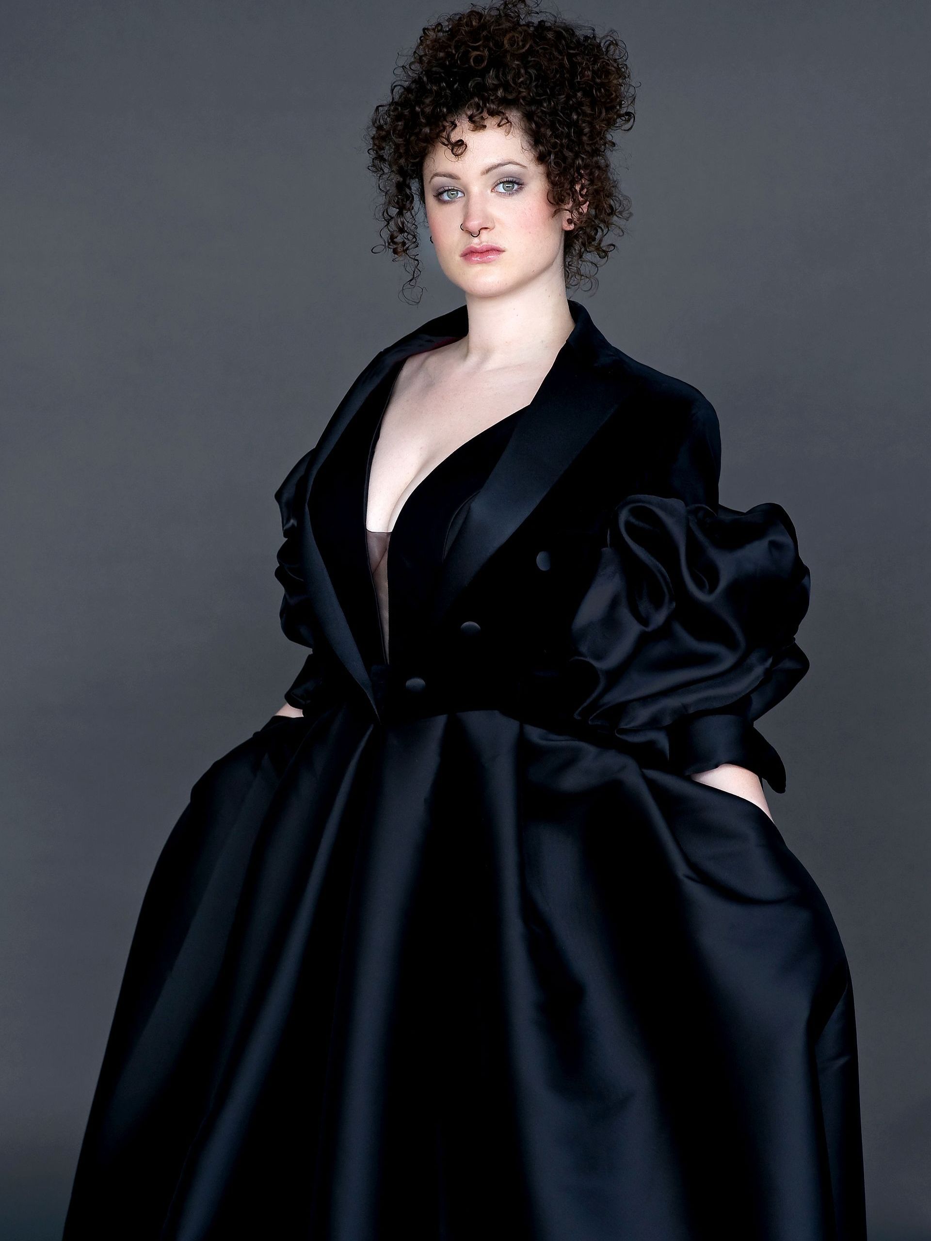 Eine Frau steht vor neutral grauem Hintergrund und trägt ein elegantes, schwarzes Kleid mit ausdrucksstarkem Design. Das Oberteil erinnert an ein tief ausgeschnittenes, doppelreihiges Jackett und geht in einen weit ausgestellten Rock über. Die voluminösen Ärmel aus glänzendem Satin verleihen dem Kleid eine dramatische, edle Wirkung. Die Frau hat lockiges, hochgestecktes Haar und blickt mit selbstbewusstem Ausdruck in die Kamera.
Dieses Kleid stammt aus der Darkdream-Kultur-Kollektion und ist erhältlich im Darkdream-Atelier für Gothic-Kleider und Steampunk-Kleidung in Gründau für Potsdam