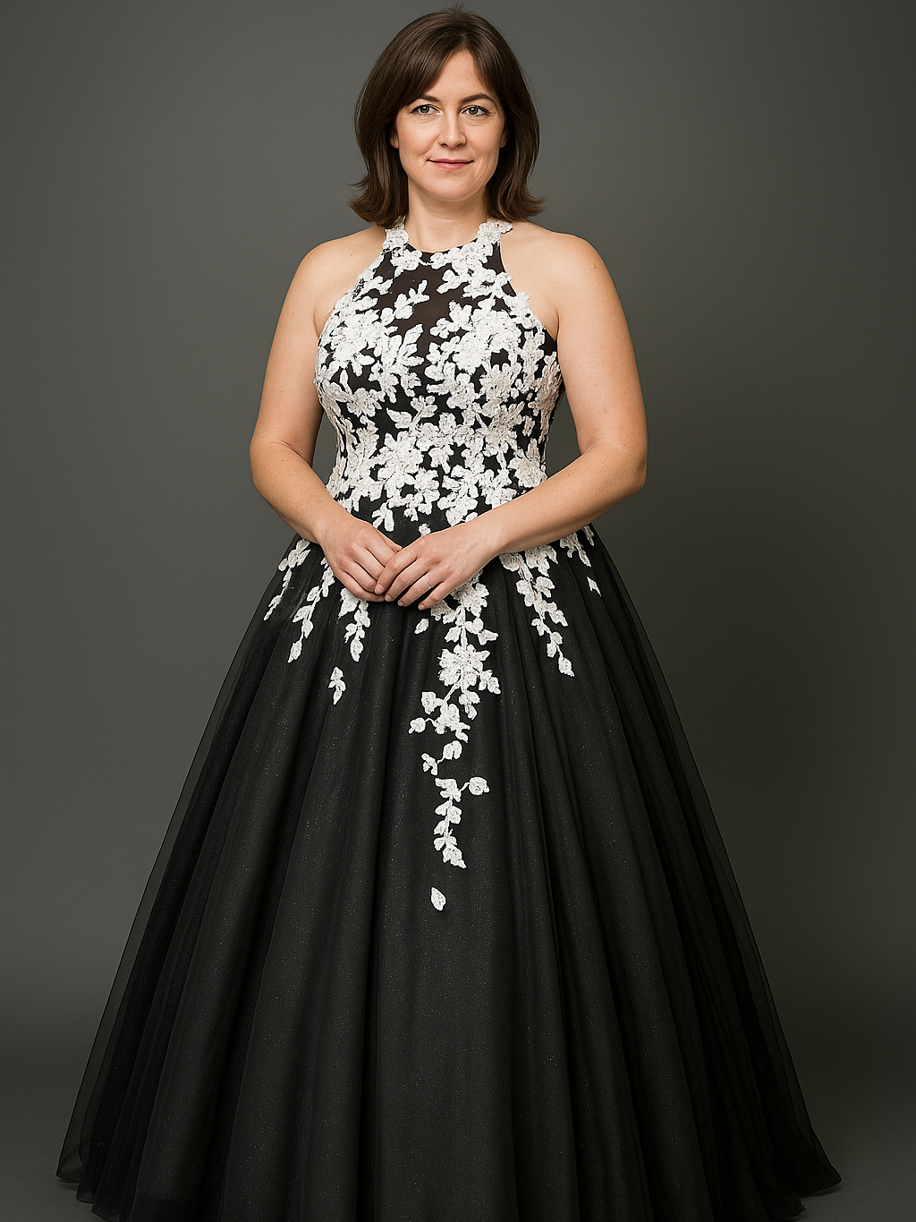 Das Bild zeigt eine Frau mittleren Alters mit schulterlangem braunem Haar vor einem neutralen, grauen Studiohintergrund. Sie trägt ein elegantes, bodenlanges Abend- oder Brautkleid im klassischen Schnitt. Das Kleid ist tiefschwarz und leicht ausgestellt mit einem weit fallenden Rock aus Tüll oder Organza. Besonders auffällig ist das Oberteil in Neckholder-Form, das aus transparentem Stoff besteht und mit kunstvoll platzierten, weißen Spitzenapplikationen in floraler Optik verziert ist. Die Applikationen ziehen sich asymmetrisch vom Oberkörper über das Kleid nach unten. Der Look wirkt romantisch und edel – eine moderne Interpretation klassischer Brautmode.
Dieses Modell gehört zur Kategorie der schwarzen Brautkleider – eine stilvolle Wahl für Bräute, die sich bewusst für Eleganz abseits des Gewöhnlichen entscheiden.
Das gezeigte schwarze Brautkleid ist erhältlich bei Mr. & Mrs. Hübner – Dark Dream Atelier in Gründau für Leipzig
