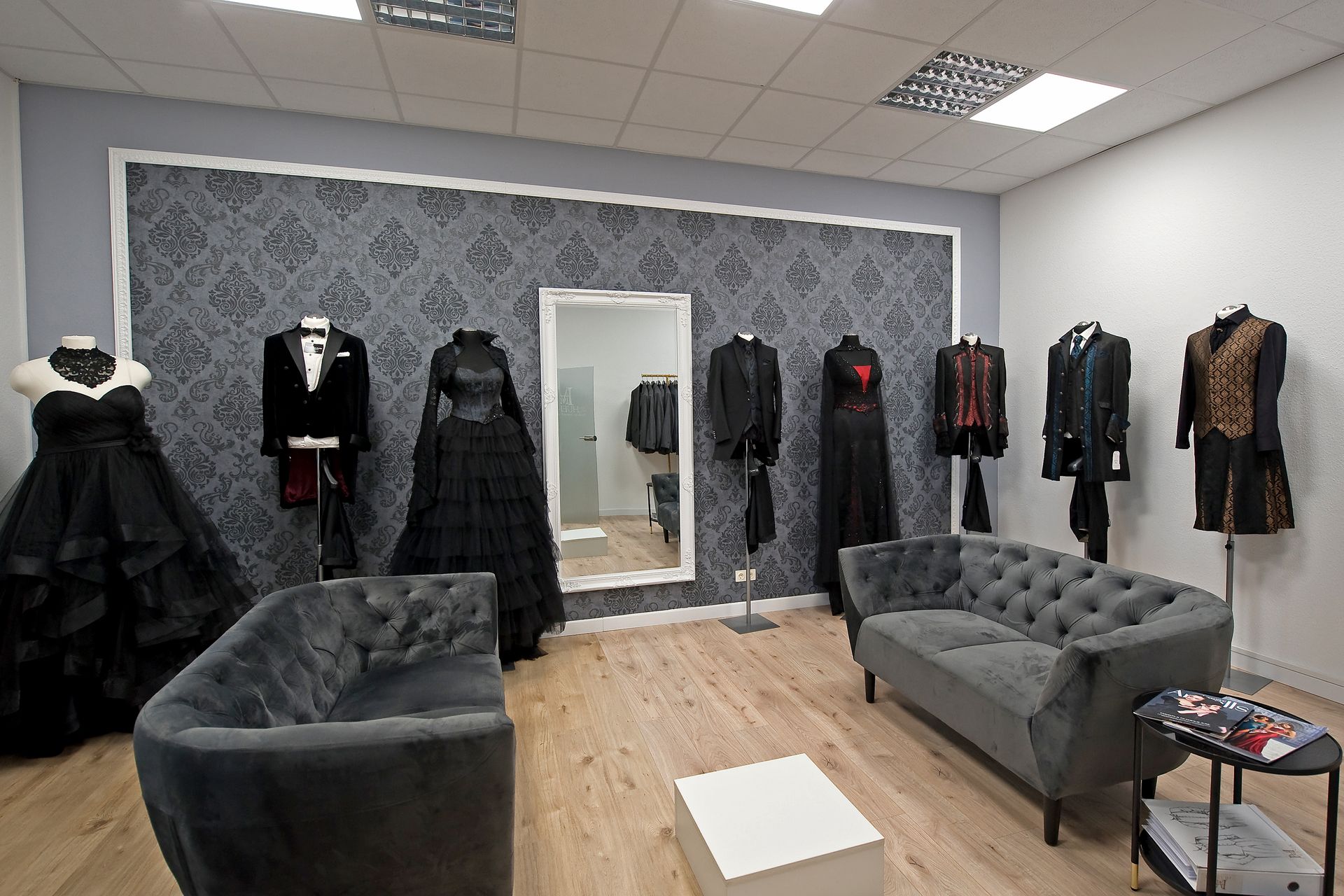 Das Bild zeigt einen stilvoll eingerichteten Raum im Dark Dream Atelier in Gründau. Im Vordergrund stehen ein dunkelgraue, Chesterfield Couchgarnitur und ein runder, niedriger Tische im Glasoptik-Stil. Auf dem Tisch Liegen Prospekte.
Im Hintergrund stehen rundherum Puppen mit den entsprechenden Outfits in verschiedenen Farben und passender Gothic-Optik. Die Farbpalette reicht von tiefem Schwarz über dunkles Rot bis zu Petrolgrün – ein visuelles Statement für Gothic-, Steampunk- und Fantasy-Mode. Die Stoffe wirken edel und hochwertig, viele Modelle besitzen auffällige Texturen wie Spitze, Satin oder Brokat.
Das Atelier strahlt eine ruhige, elegante Atmosphäre aus und bietet den perfekten Rahmen für persönliche Beratung und Anproben. Das Bild unterstreicht den exklusiven, außergewöhnlichen Stil der Marke Dark Dream Collection.