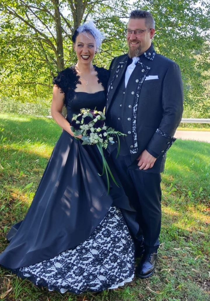 Gothic-Brautpaar im Black & White Stil: Die Braut trägt ein schwarzes A-Linien-Kleid mit weißer Spitzenlage im Unterrock, der Bräutigam einen schwarzen Gehrock mit schwarz-weißem Besatz an Revers und Manschetten – aufgenommen im Grünen bei Sonnenlicht.