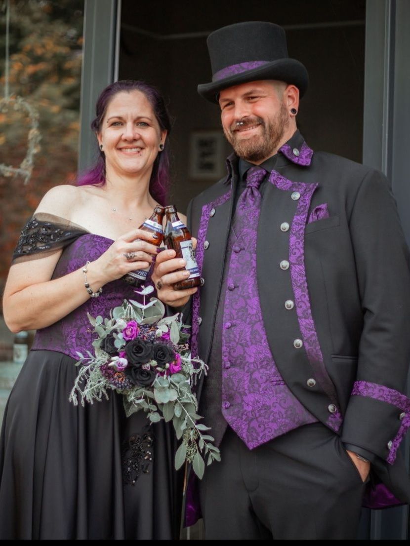 Das Bild zeigt ein fröhliches Paar in festlicher Gothic- und Steampunk-inspirierter Hochzeitskleidung. Beide lächeln in die Kamera und stoßen mit kleinen Bierflaschen an. Sie stehen nebeneinander im Außenbereich vor einer Glastür, die den Blick auf eine herbstliche Umgebung freigibt.
Die Frau auf der linken Seite trägt ein schulterfreies Kleid in Schwarz und kräftigem Lila. Das Oberteil besteht aus einer violetten Brokatkorsage mit floralen Mustern und schwarzen Spitzenapplikationen an den Schultern. Ihr Rock ist lang und schwarz. In der linken Hand hält sie einen Hochzeitsstrauß aus schwarzen und violetten Rosen sowie silbrig-grünem Blattwerk – passend zur Farbgebung ihres Outfits. Ihre Haare sind violett gefärbt und offen getragen.
Der Mann auf der rechten Seite trägt ein schwarzes Hochzeitsensemble mit auffälligen lilafarbenen Brokat-Details: eine gemusterte Weste, ein Einstecktuch, eine Krawatte und dekorative Besätze am Gehrock. Sein Outfit wird durch einen klassischen Zylinderhut mit lilafarbenem Band ergänzt. Er hat ein gepflegtes Bartstyling, einen Nasenring und mehrere Ohrpiercings. Beide wirken gut gelaunt, natürlich und verbunden.
Dieses Bild zeigt eine lebendige, individuelle Interpretation alternativer Hochzeitsmode – festlich, verspielt und farblich perfekt abgestimmt.
Die abgebildeten Gothic-Hochzeitsoutfits sind erhältlich bei Mr. & Mrs. Hübner – Dark Dream Atelier in Gründau für Berlin