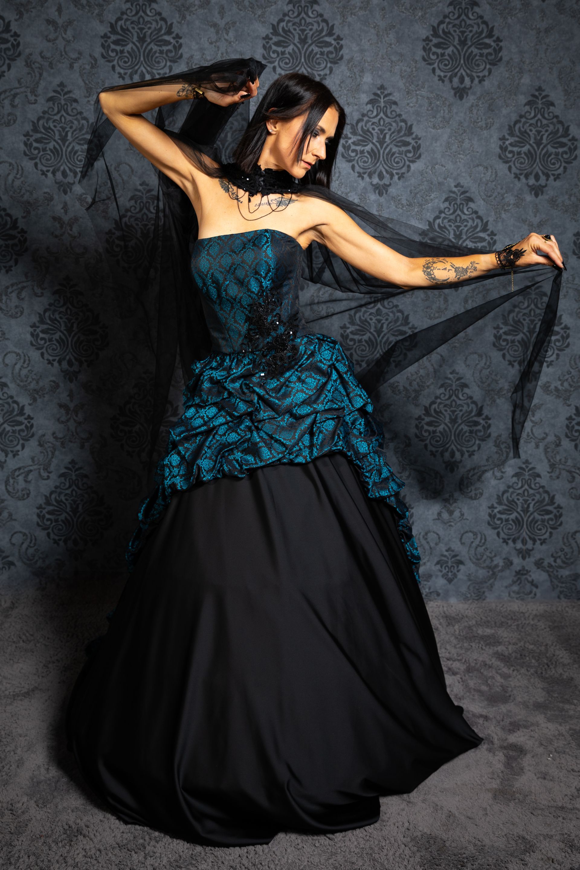 Eine Frau steht seitlich vor einer alten viktorianischen Tapet. Sie trägt ein schulterfreies Kleid im Victorian Art Style mit einem weit ausfallenden schwarzen Rock. Das Oberteil und der obere Rockteil sind in tiefem Petrol mit schwarzem Brokatmuster gestaltet, darüber liegt ein leichter schwarzer Tüllschleier, den sie mit beiden Händen seitlich ausbreitet. Ihr langes, schwarzes Haar fällt glatt über die Schultern. Sie trägt ein auffälliges schwarzes Halsband mit Schmuckelementen sowie dunkles, ausdrucksstarkes Make-up. Auf ihren Armen sind tätowierte Motive sichtbar. Die Szenerie wirkt dramatisch, geheimnisvoll und elegant.
Dieses Outfit ist im Gothic-Style gehalten und erhältlich im Dark Dream Atelier für Gothic- und Steampunk-Mode in Gründau.