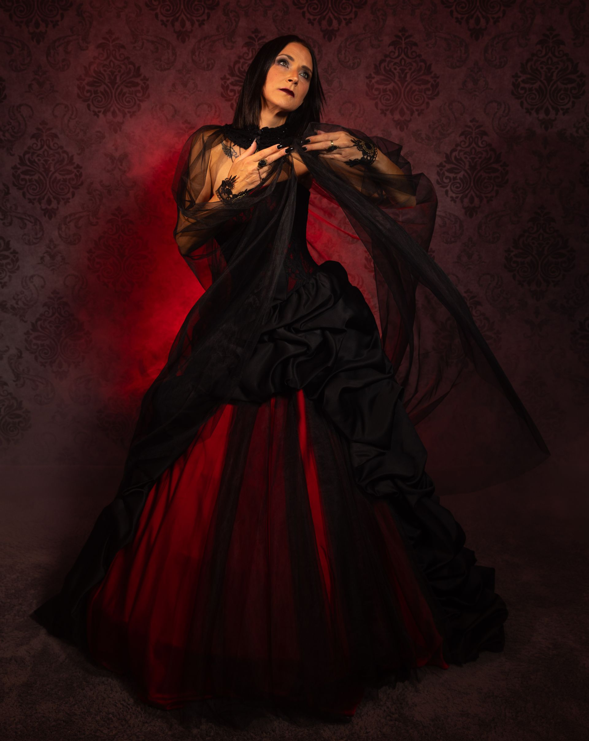 Frau in einem dramatischen schwarz-roten Gothic-Kleid mit transparentem Tüll und aufwändigen Drapierungen, posiert vor einer dunklen, ornamentalen Tapete. Roter Lichteffekt im Hintergrund verstärkt die mystische und elegante Atmosphäre.
Erhältlich bei Mr. & Mrs. Huebner – Dark Dream Atelier in Gründau, in der Kategorie Schwarze Brautkleider.