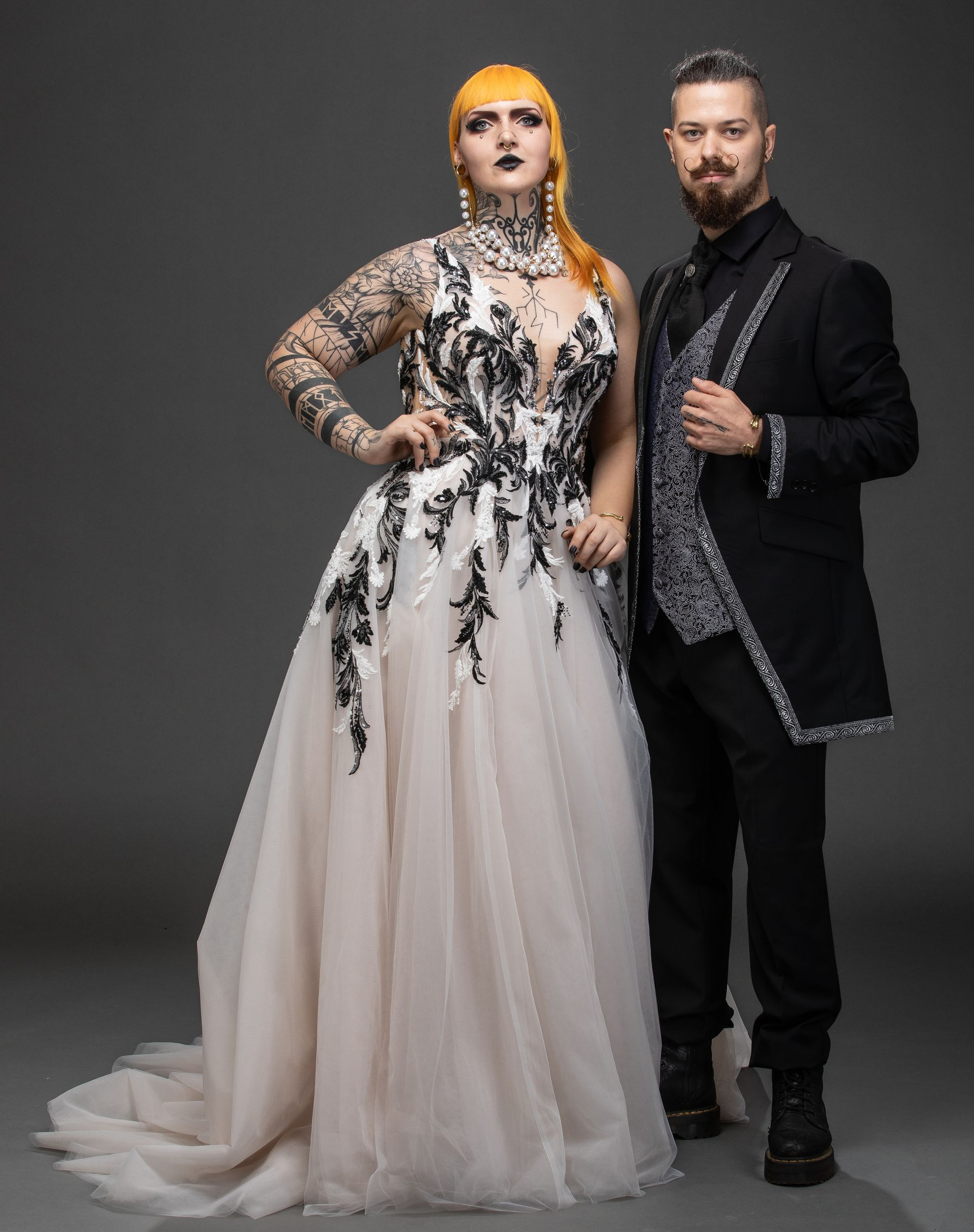 Das Bild zeigt ein elegant gekleidetes Paar vor einem dunklen, einfarbigen Hintergrund. Links im Bild steht ein Mann mit kurzem Haar, Bart und Brille. Er trägt einen edlen, schwarzen Anzug mit schwarzem Hemd und schwarzer Krawatte. Seine Hände ruhen locker in den Hosentaschen, sein Blick ist ruhig und freundlich.
Rechts neben ihm steht eine Frau mit dunklem, leicht gewelltem Haar, das offen über die Schultern fällt. Sie trägt ein glamouröses, bodenlanges Abendkleid in Schwarz. Das Kleid ist im Oberkörperbereich figurbetont und mit floralen Verzierungen und Glitzerelementen bestickt. Die langen, transparenten Ärmel sind aus zartem Tüll gearbeitet. Der Rock ist leicht ausgestellt und wirkt durch den changierenden Stoff, den Glitzer und die Spitzenapplikationen besonders edel. Der Look verbindet romantische Eleganz mit moderner Zurückhaltung.
Die Gesamtwirkung des Bildes ist stilvoll, hochwertig und harmonisch – typisch für den Modestil Black Edelschick, der dezente Raffinesse und dunkle Ästhetik verbindet.
Das gezeigte Outfit stammt aus der Kollektion „Black Edelschick“ und ist erhältlich bei Mr. & Mrs. Huebner – Dark Dream Atelier in Gründau