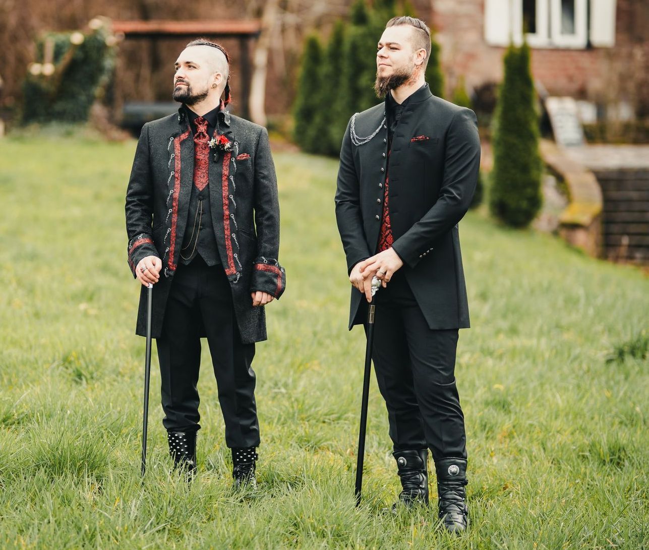 Das Bild zeigt zwei Männer in festlicher, alternativer Herrenmode mit starkem Gothic- und viktorianischem Einfluss. Sie stehen nebeneinander auf einer grünen Wiese im Außenbereich vor einem historischen Gebäude mit Backsteinfassade. Beide Männer tragen schwarze Outfits mit auffälligen roten Akzenten und halten jeweils einen Spazierstock – was ihrem Auftritt eine aristokratische Note verleiht.
Der Mann auf der linken Seite hat einen seitlich rasierten Haarschnitt mit dunklem Oberhaar, trägt einen schwarzen Anzug mit roten Brokat-Details am Revers, an den Manschetten und an der Knopfleiste. Unter dem Jackett ist eine rote gemusterte Weste zu sehen. Seine Schuhe sind schwarz und mit Metallakzenten versehen. Am Revers trägt er eine rote Ansteckblume.
Der Mann auf der rechten Seite ist größer, hat einen Vollbart und trägt einen hochgeschlossenen Gehrock mit asymmetrischem Revers, Schulterkette und roten Details, die nur dezent hervorblitzen. Auch sein Spazierstock passt farblich und stilistisch zum Outfit. Seine Stiefel sind geschnürt und mit dekorativen Schnallen versehen.
Beide wirken selbstbewusst, elegant und kraftvoll – ihr Look kombiniert historische Elemente mit moderner Szeneästhetik und ist ein Beispiel für exklusive Gothic-Herrenanzüge mit Steampunk- und Dark-Style-Charakter.
Die gezeigten Herrenoutfits sind erhältlich bei Mr. & Mrs. Hübner – Dark Dream Atelier in Gründau für Rostock