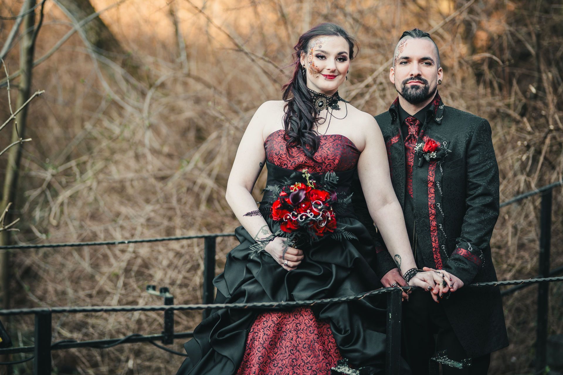 Ein Paar steht im Freien vor einem herbstlich wirkenden Hintergrund mit kahlen Ästen. Die Frau trägt ein aufwendig gestaltetes, schulterfreies Brautkleid im Gothic-Stil. Das Oberteil und ein Teil des Rockes sind in dunklem Rot mit schwarzem Brokatmuster gehalten, der restliche Rock ist schwarz und voluminös. Sie hält einen Brautstrauß aus roten und schwarzen Blumen mit Schmuckelementen. Ihr langes, dunkles Haar ist teils hochgesteckt, teils offen und fällt über die Schulter. Sie trägt auffälliges, dunkles Make-up mit roten und schwarzen Akzenten sowie ein schwarzes Halsband mit Ornamenten. Der Mann steht rechts neben ihr, trägt einen schwarzen Gehrock mit roten Brokatverzierungen an den Revers und Ärmelabschlüssen. Er hat dunkles, zurückgekämmtes Haar mit rasierten Seiten und einen gepflegten Bart. Beide halten sich an den Händen und schauen in die Kamera. Die Stimmung wirkt romantisch, stilvoll und leicht mystisch.
Dieses Outfit ist im Gothic-Style gehalten und erhältlich im Dark Dream Atelier für Gothic- und Steampunk-Mode in Gründau für Dresden
