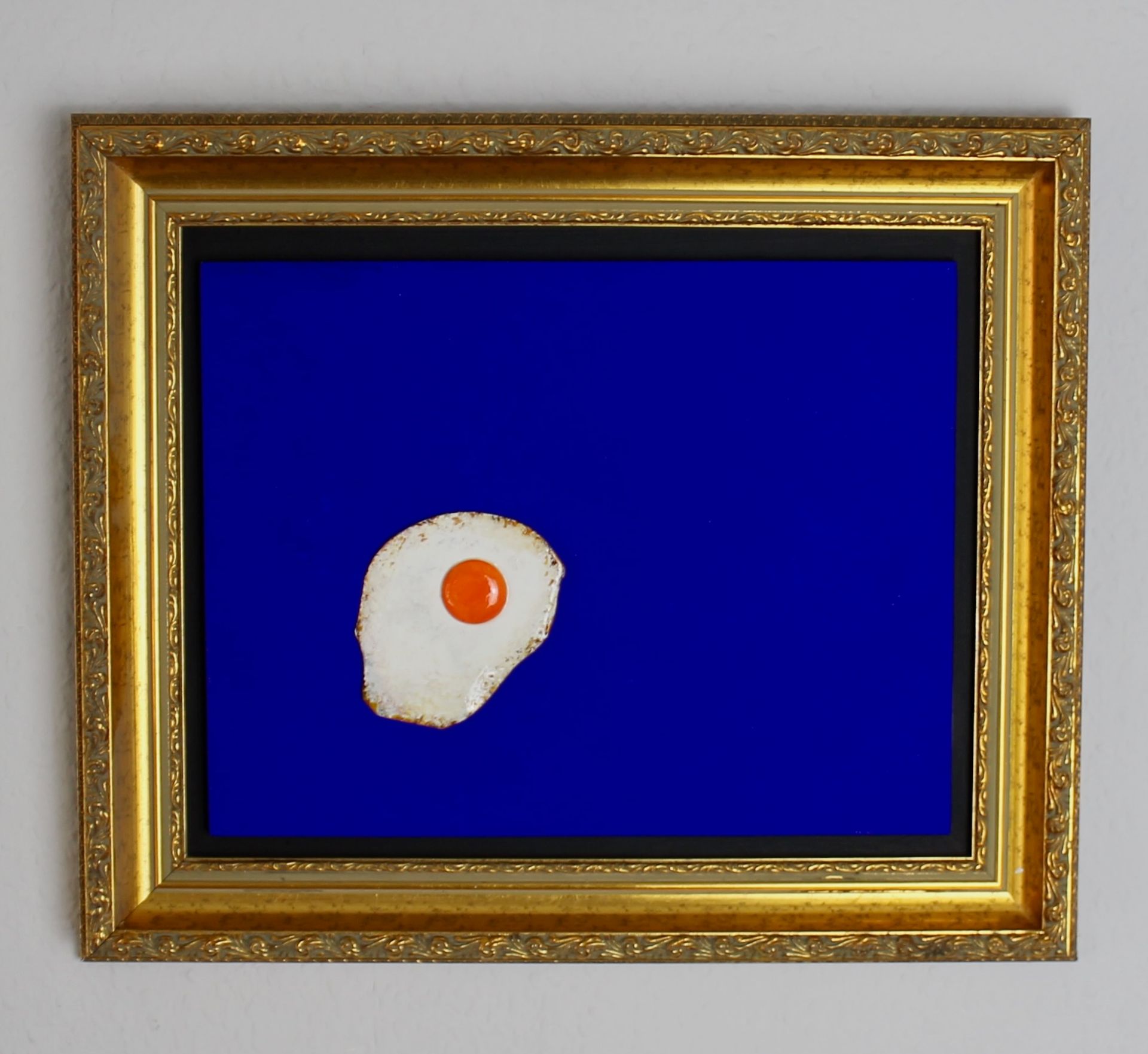 Das Bild zeigt ein Spieglei auf einem blauen Grund im Goldrahmen. Ein beliebtes Motiv von Bildhauer Oliver Pohl aus Haste im Schaumburger Land in der Nähe von Bad Nenndorf.