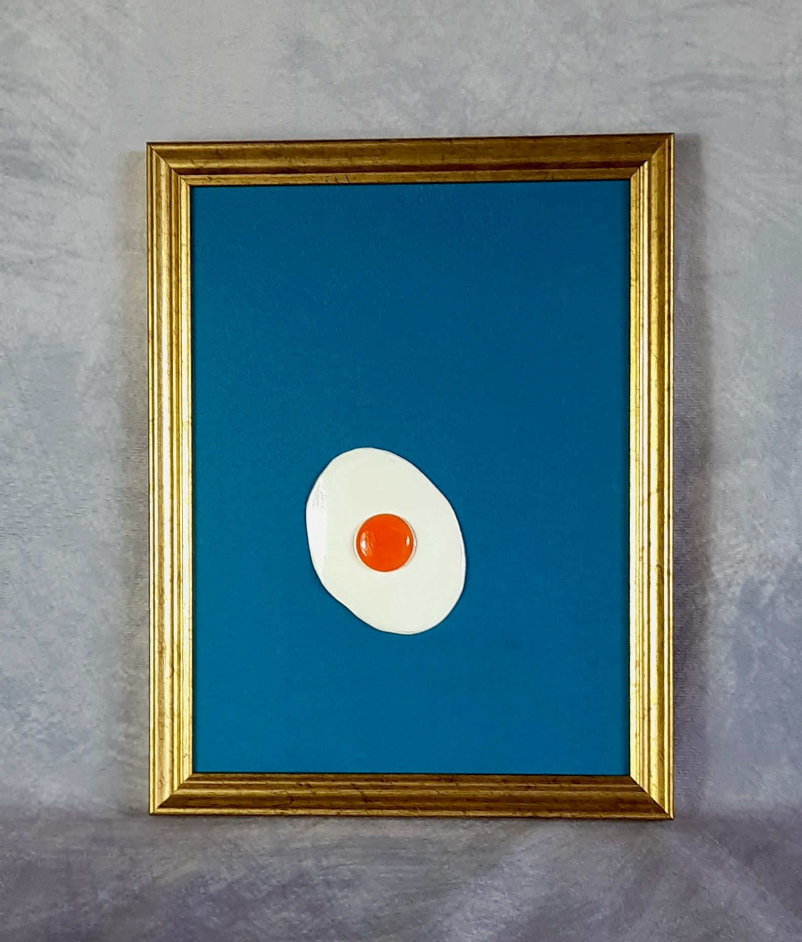 Plastisches Pop Art Spiegelei auf türkisem Grund in einem goldenen Rahmen.