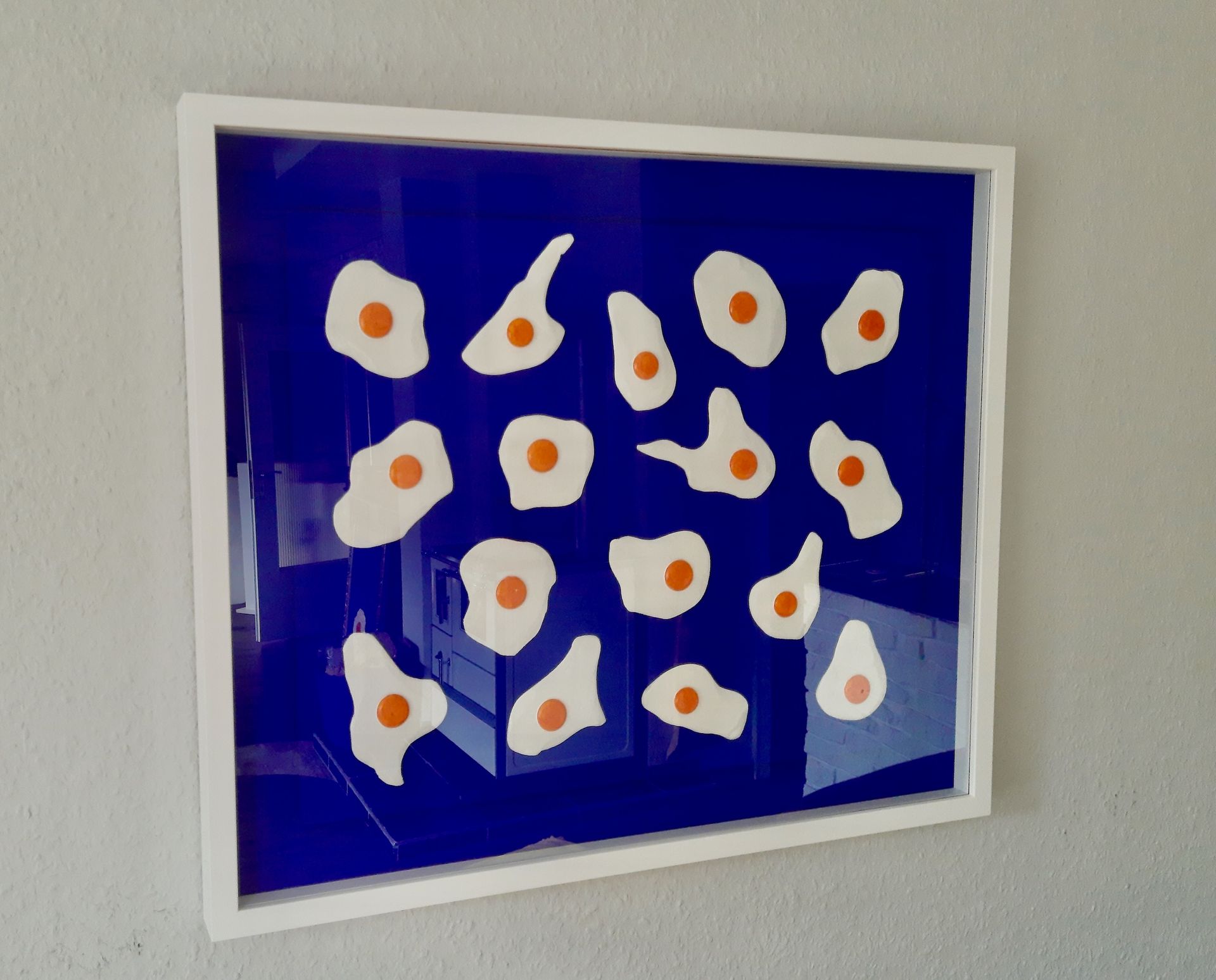 Glasrahmen mit 15 plastischen Spiegeleiern auf ultramarinem Grund.