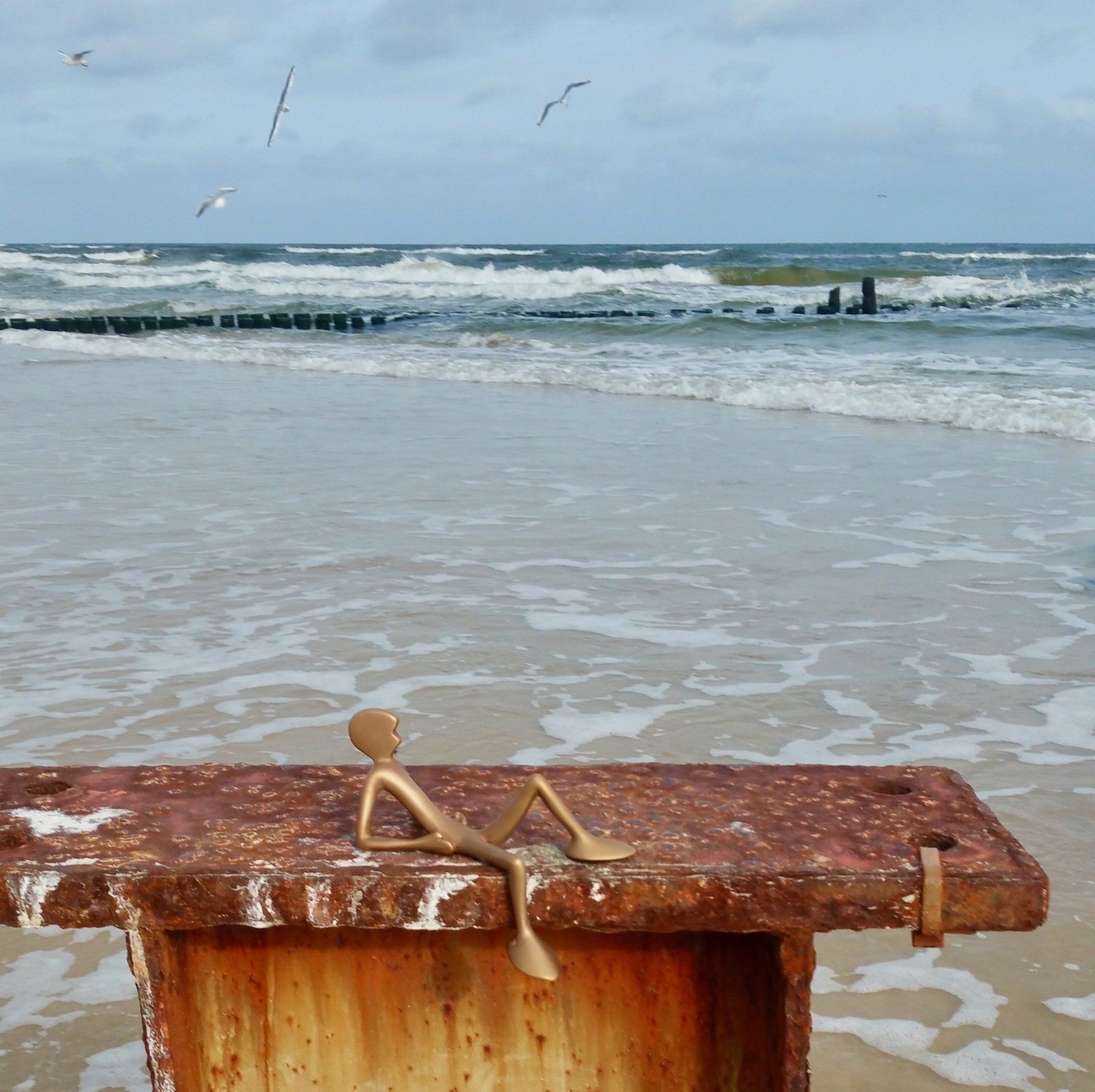 Das Bild zeigt den Bronzefriend No 3 von Bildhauer Oliver Pohl. Er liegt ganz lässig auf einem Metallpoller am Strand von Heringsdorf. Im Hintergrund ist die Ostsee zu sehen.