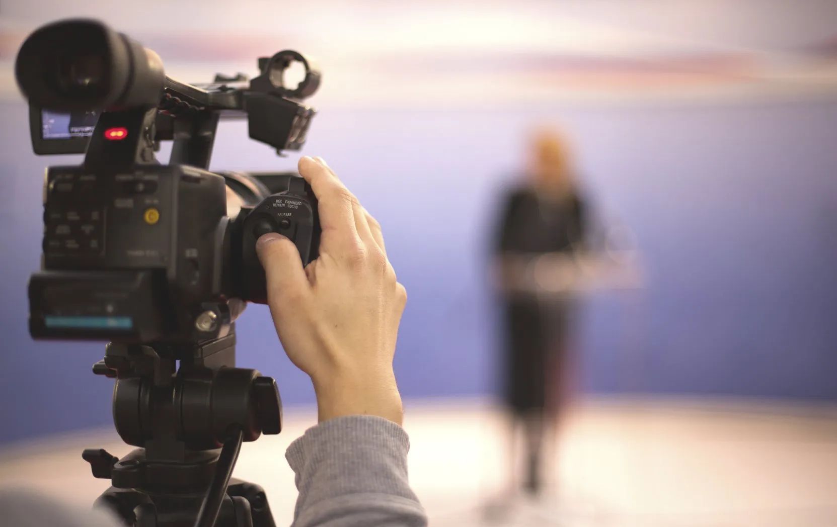 Videokamera filmt Person Rückseite einer Videokamera mit Hand am Auslöser. Vor der Kamera unscharf eine Person.