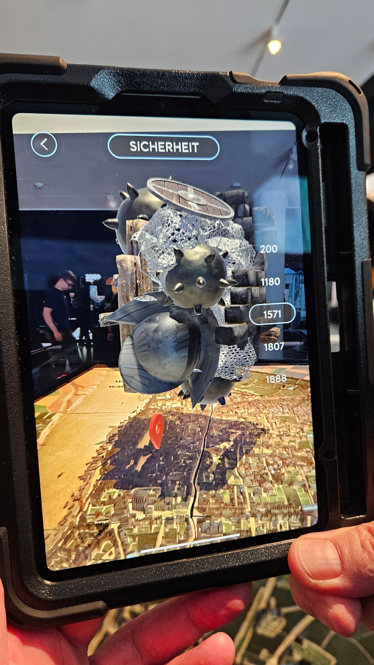 Der Schirm eines bildfüllenden Tablets zeigt im Hintergrund ein Stadtmodell. Davor ist eingeblendet ein seltsames kompaktes Objekt das metallisch glänzt und aus verschiedenen Kugelformen besteht. Auf manchen sind spitze Zacken. Rechts eine Zeitleiste mit Jahreszahlen - 1571 weiß umrandet.