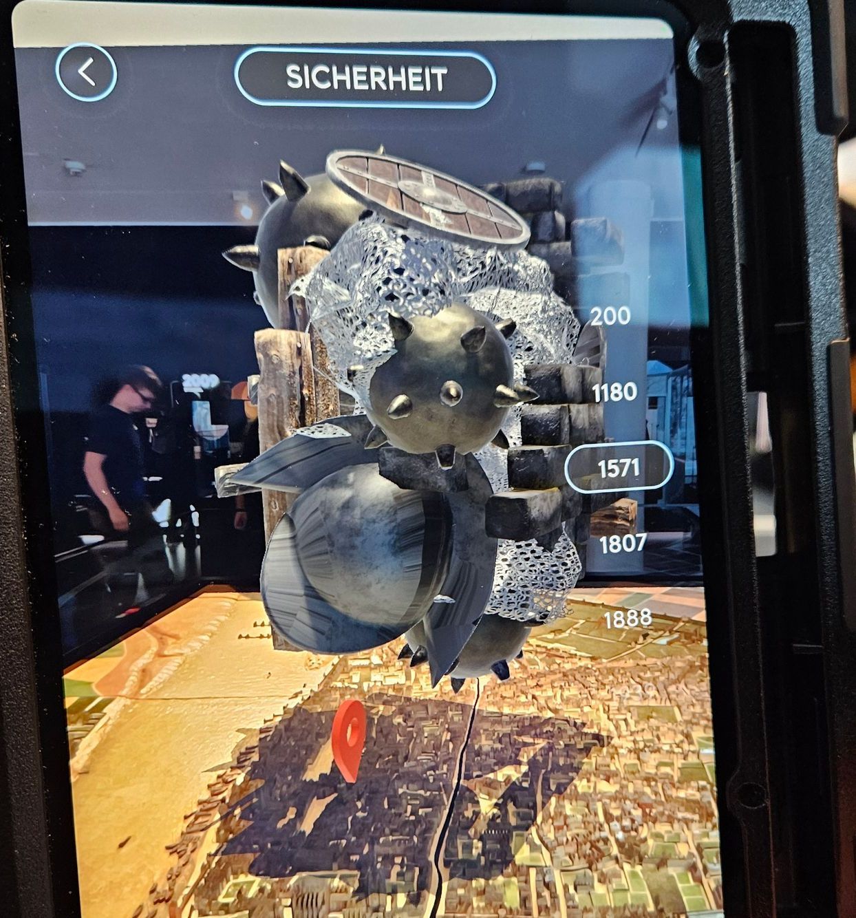 Der Schirm eines bildfüllenden Tablets zeigt im Hintergrund ein Stadtmodell. Davor ist eingeblendet ein seltsames kompaktes Objekt das metallisch glänzt und aus verschiedenen Kugelformen besteht. Auf manchen sind spitze Zacken. Rechts eine Zeitleiste mit Jahreszahlen - 1571 weiß umrandet.