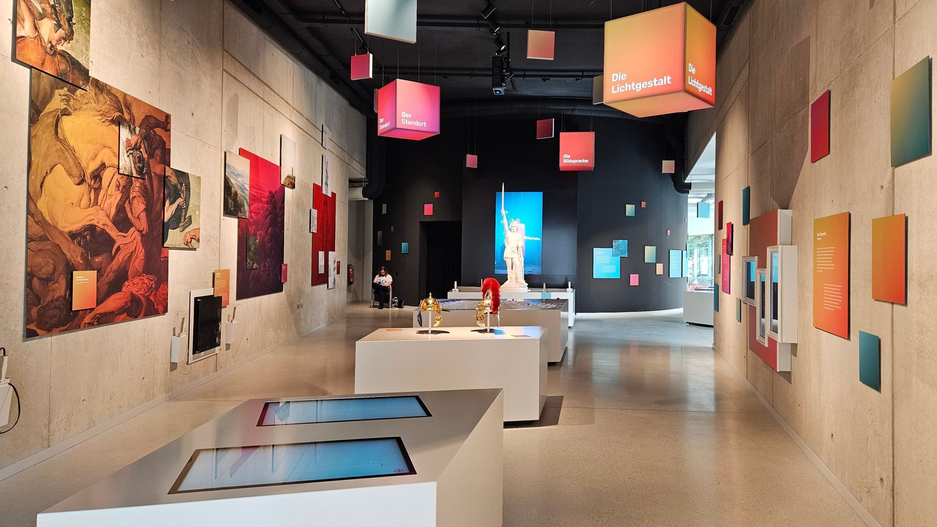 Ein Ausstellungsraum. Im Vordergrund ein weißes Möbel mit zwei schräg angeordneten großen Monitoren. Anden Wänden verschieden große farbige Tafeln Von der Decke hängen rote und orangene Würfel mit weißen Texten: Die Lichtgestalt, Der Standort.