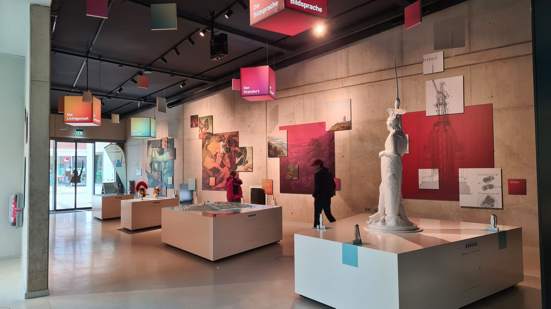 Ein Ausstellungsraum mit mehreren Podesten. Darauf vorne eine Statue in Weiß. Weitere Objekte auf den Podesten dahinter. An der Wand Bilder auf roten rechteckigen Hintergrundflächen.