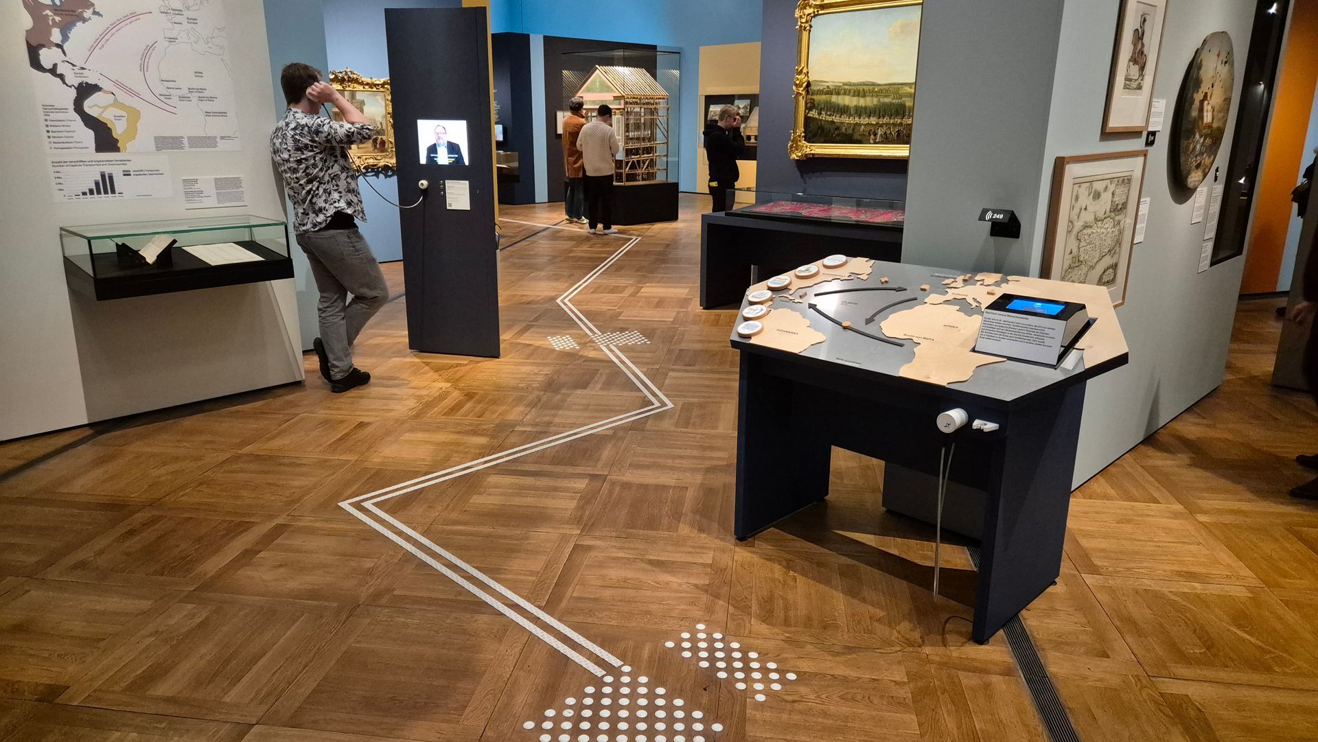 Ein Ausstellungsraum mit Stelen, Vitrinen und im Vordergrund ein sechseckiger Tisch. Darauf eine Weltkarte im Relief, gelb auf grauem Grund. Links flache runde Objekte und rechts ein offenbar drehbares Objekt mit mehreren Flächen: Darauf vorne ein Text und oben ein Bildschirm. Vorne am Tisch ein Ohrhörer und eine Klemmvorrichtung. Auf dem Boden weiße Leitstreifen mit einem Punktefeld vor dem Tisch.