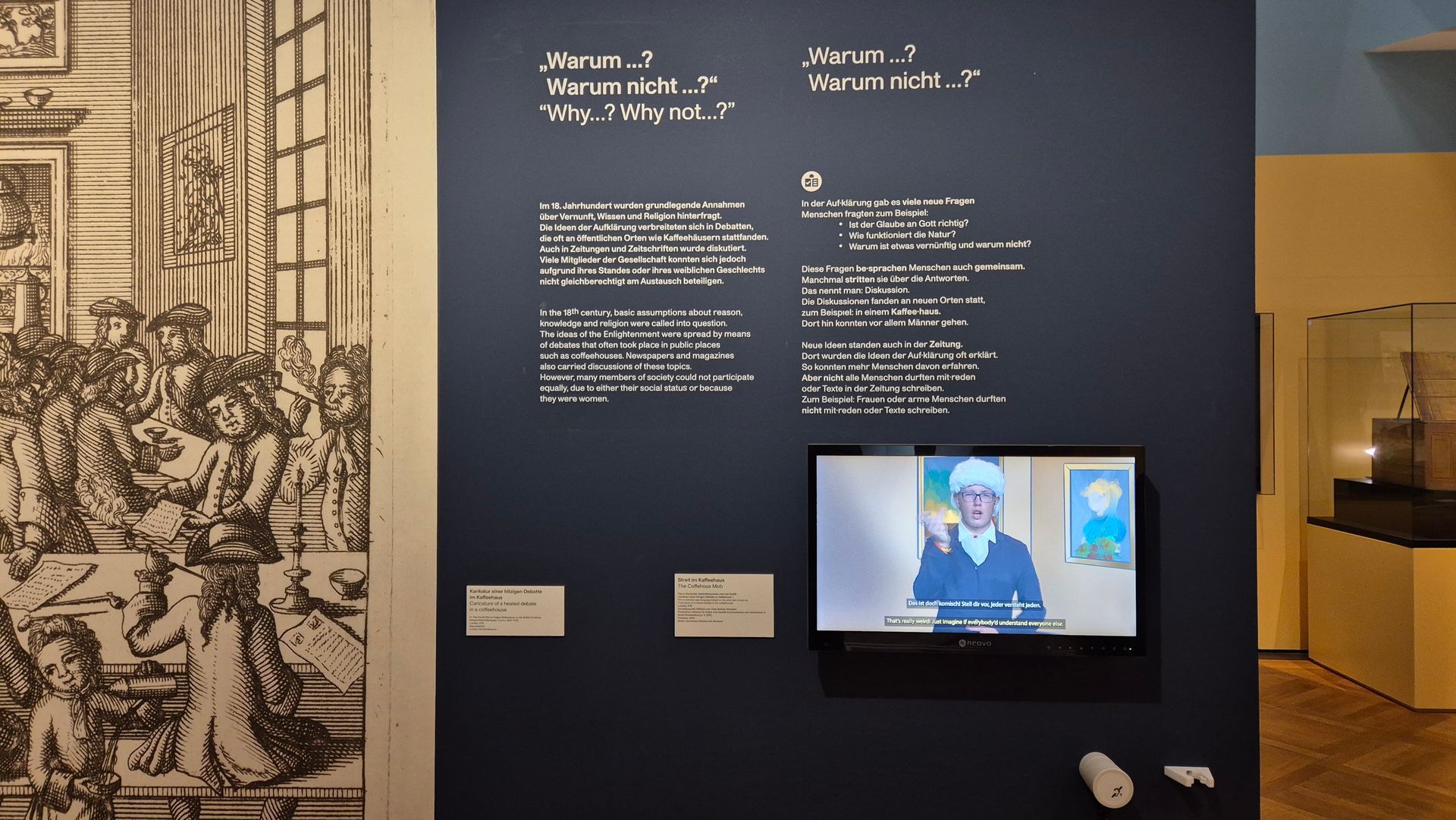 Eine Ausstellungswand mit weißen Texten auf schwarzem Grund. Rechts ein kleiner Monitor mit einer Person.