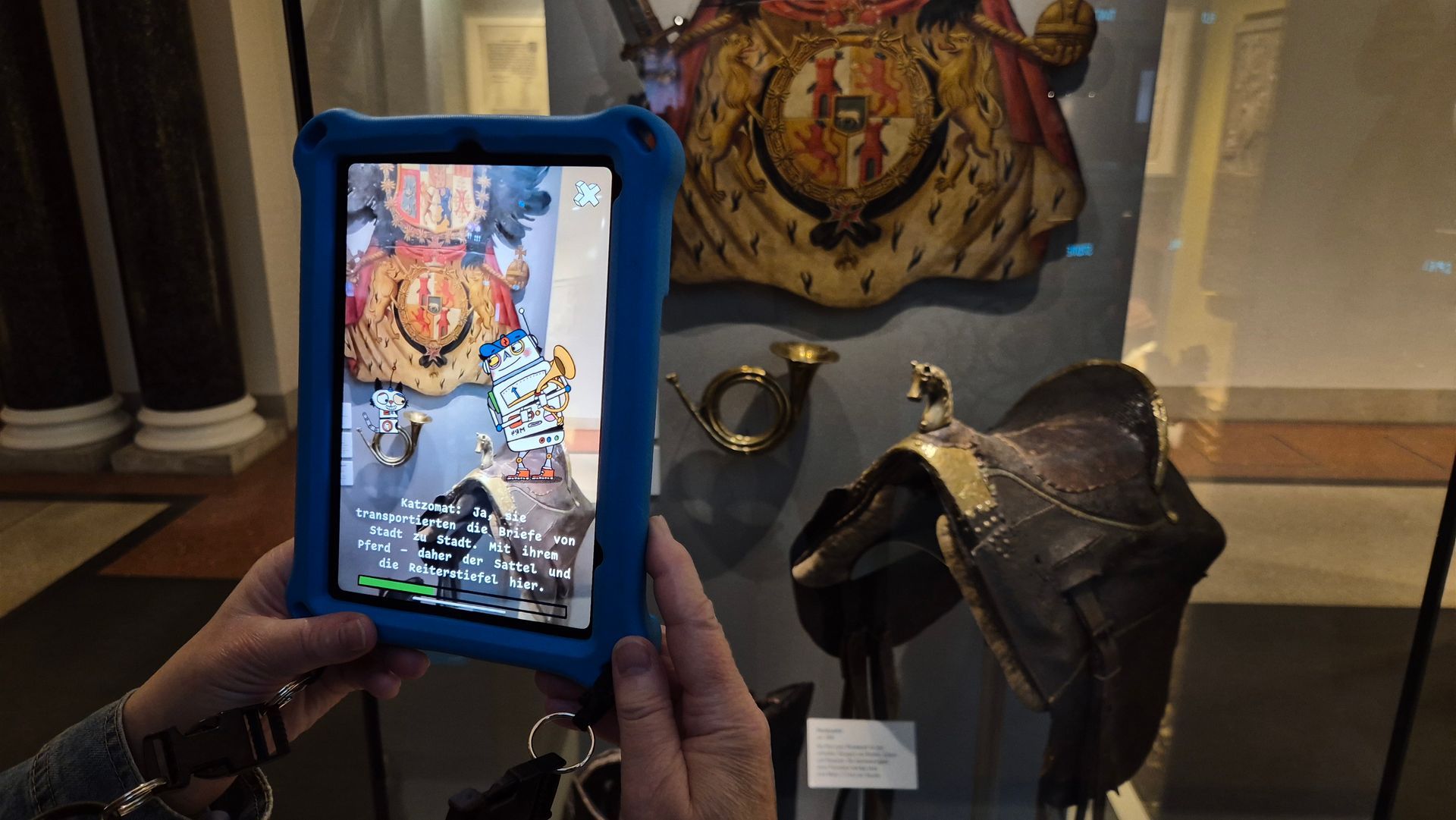 Eine Hand hält ein Tablet vor eine Vitrine mit einem Sattel, einem großen Stoffwappen und einem kleinen Posthorn. Auf dem Monitor sitzt die kleine Roboterkatze auf dem Posthorn. Redchts auf dem Sattel steht der kastige Blechroboter und hält ein Posthorn in der Hand. Darunter steht ein Text.