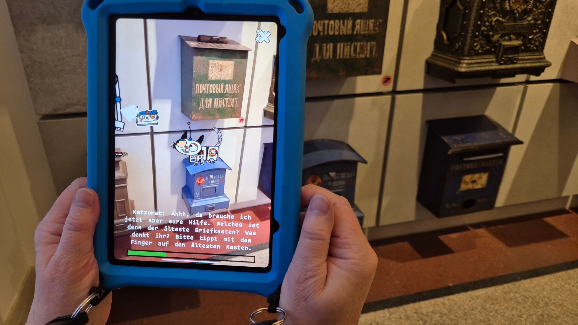 Eine Hand hält ein Tablet vor eine Wand voller alter Biefkästen. Auf dem Monitor sitzt die kleine Roboterkatze auf einem blauen Kasten. Darüber ein sehr alter olivgrüner Briefkasten mit gelber Schrift. Unten steht ein Text.