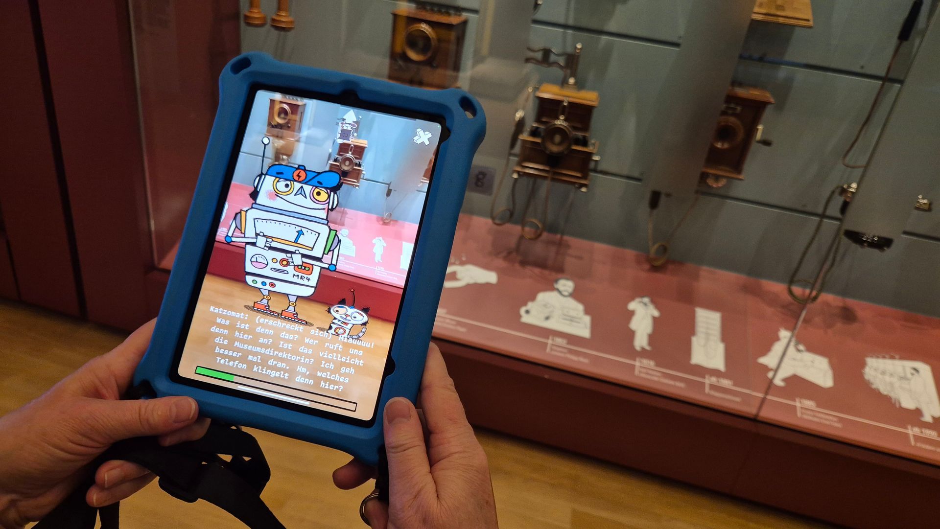 Eine Hand hält ein Tablet vor eine Glasvitrine mit alten Telefonen. Auf dem Bildschirm stehen zwei gezeichnete Roboter vor dieser Vitrine: Ein kastenförmiger lustiger Kerl mit blauem Käppi und eine kleinere Roboterkatze. Darunter einText.