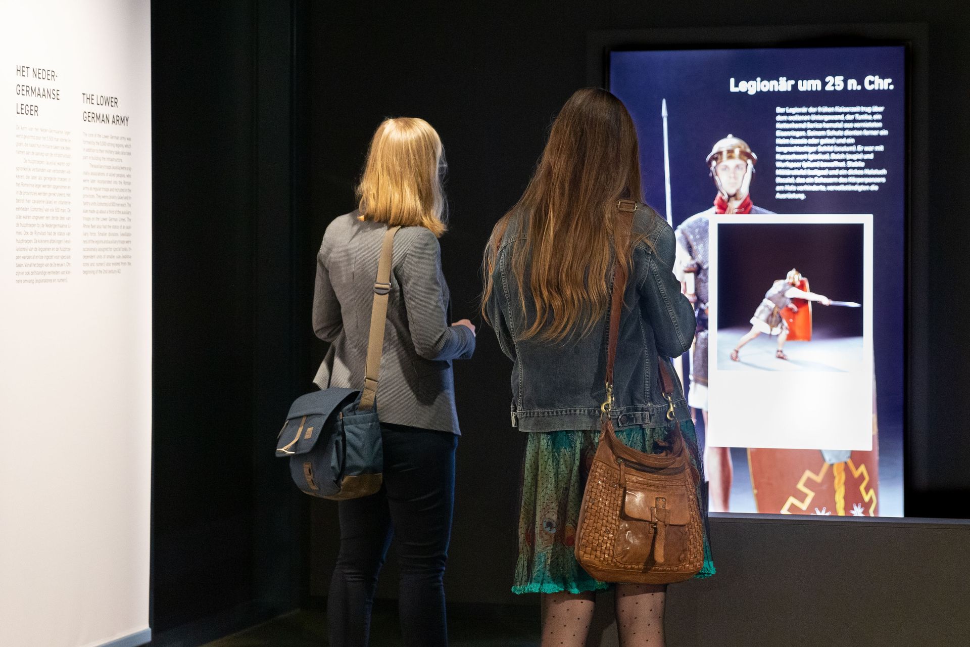 Zwei Frauen stehen vor einem hochformatigen Monitor mit römischem Legionär.