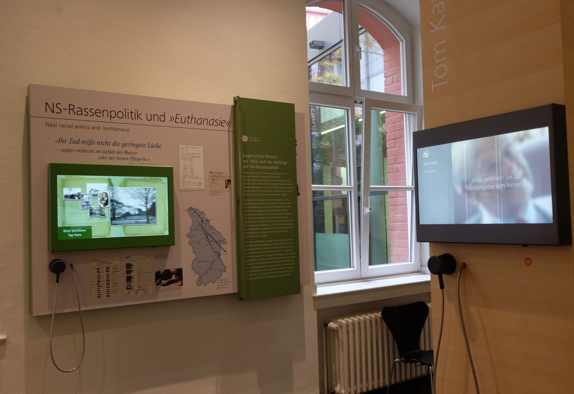 Ein Ausstellungsraum, auf der Wand links ein Monitor mit Bildern und Texten, rechts ein altes Fenster, das nach drauße geht und im Vordergrund eine Wand mit einem Monitor mit Einhandhörer. Darauf das Gesicht einer Person.