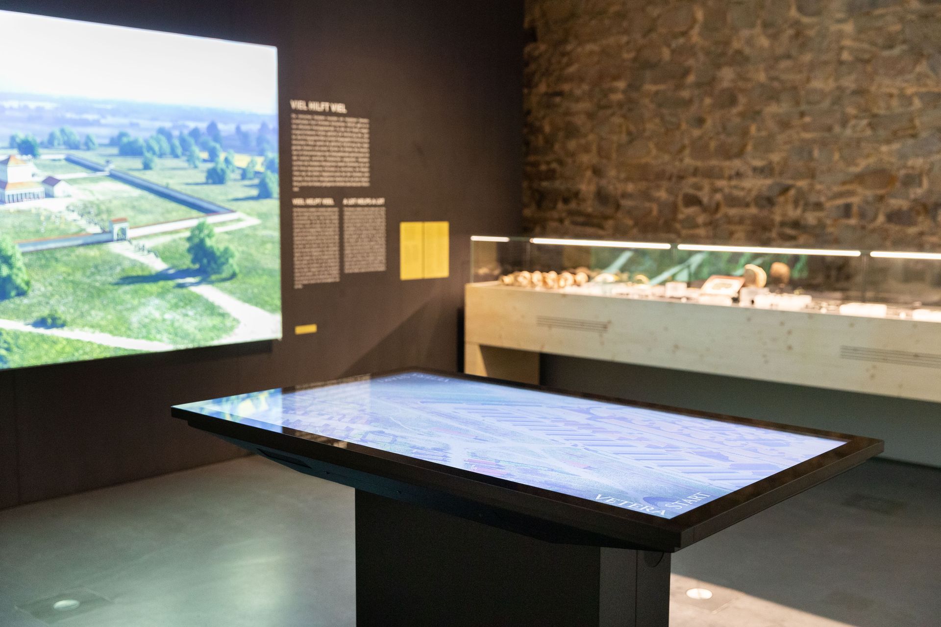 Ausstellungsraum mit einem leuchtenden Monitortisch im Vordergrund. Hinten eine flache Vitrine mit Objekten.