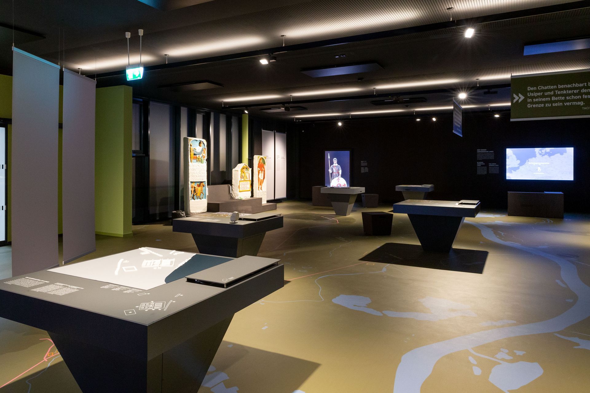 Raumansicht der Ausstellung. Graue Tische mit Texten und Objekten verteilt. Im Hintergrund ein großer Monitor mit einem römischen Soldaten.