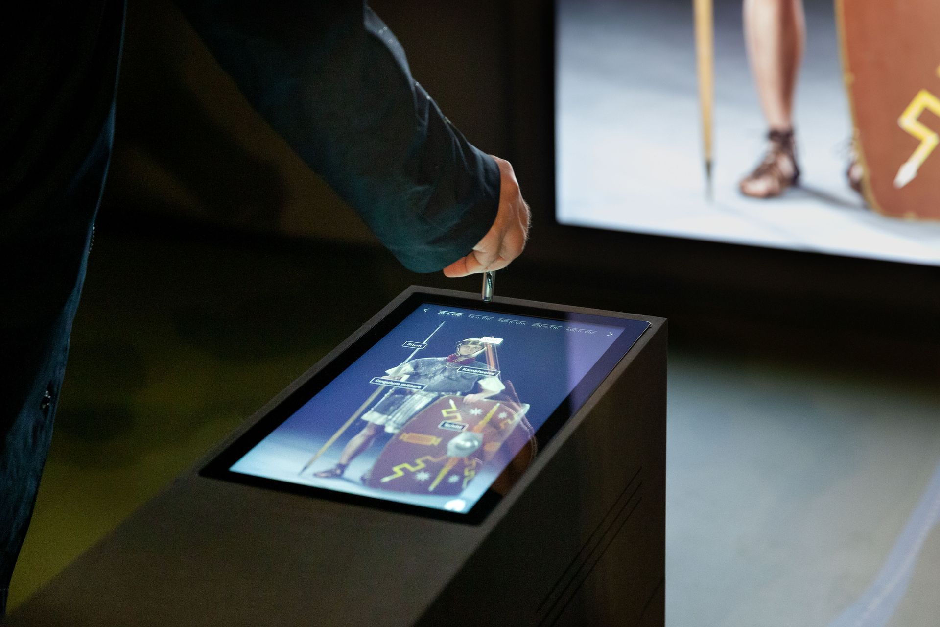 Schräg eingebauter Touchmonitor. Darauf ein römischer Soldat in Rüstung. Eine Hand tippt mit einem Stift auf das Glas. Im Hintergrund angeschnitten ein großer Bildschirm. Darauf angeschnitten ein Bein eines Soldaten.