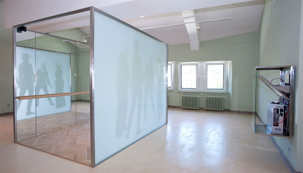 Ein großer begehbarer Quader, der an der rechten Seite eine Milchglasscheibe hat, auf die die Schatten tanzender Personen projiziert werden.
