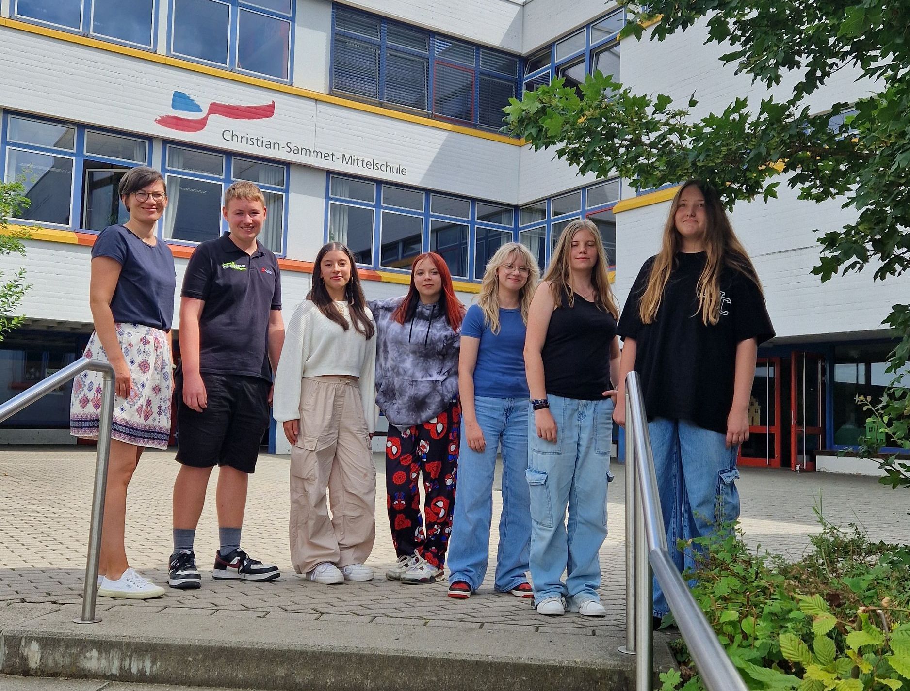 Frau mit Schüler und Schülerinnen vor Schulgebäude.