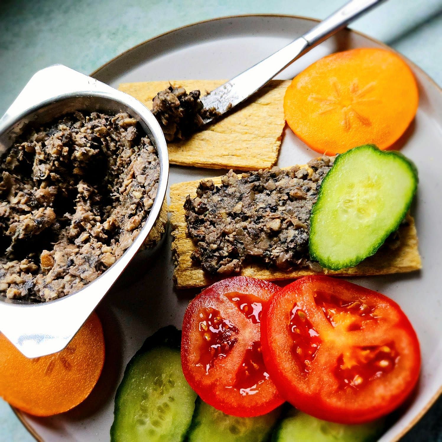 vegan bean and walnut pâté in a silver dish spread on crispbread with fresh tomato slices, sliced cucumber, and a slice of persimmon.