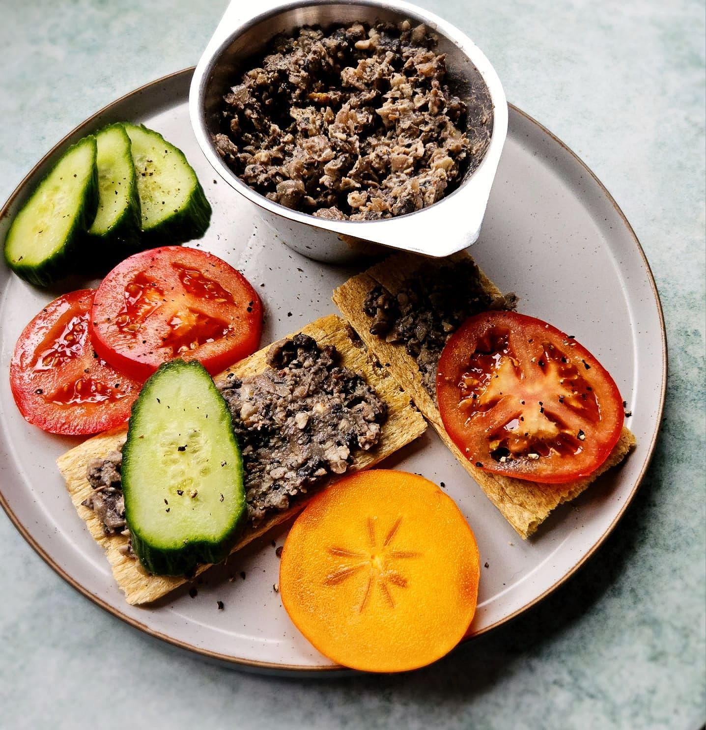 vegan bean and walnut pâté in a silver dish spread on crispbread with fresh tomato slices, sliced cucumber, and a slice of persimmon.