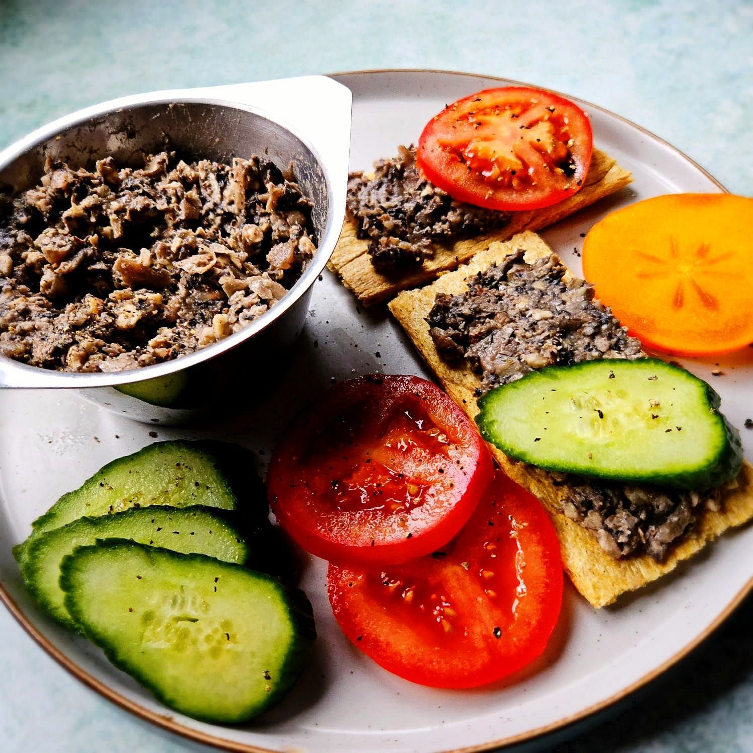 vegan bean and walnut pâté in a silver dish spread on crispbread with fresh tomato slices, sliced cucumber, and a slice of persimmon.