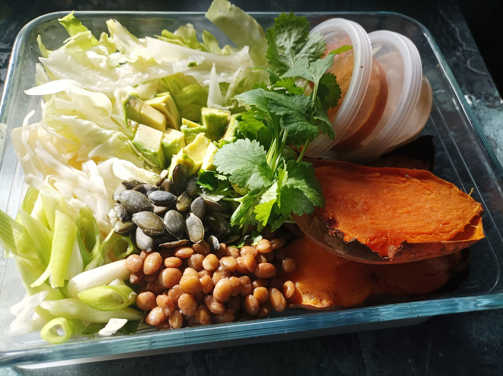 buddha bowl containing lentils, sweet potato, salad and chopped avocado in a glass dish with a dark and light background