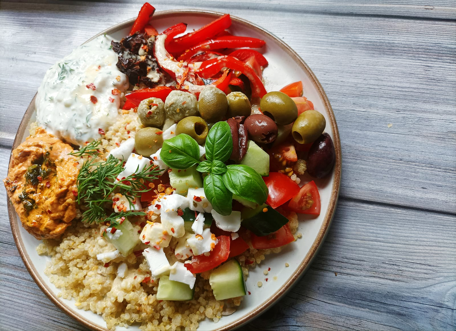 a bowl of quinoa salad with veggies, vegan feta, fresh basil, hummus and tzatziki