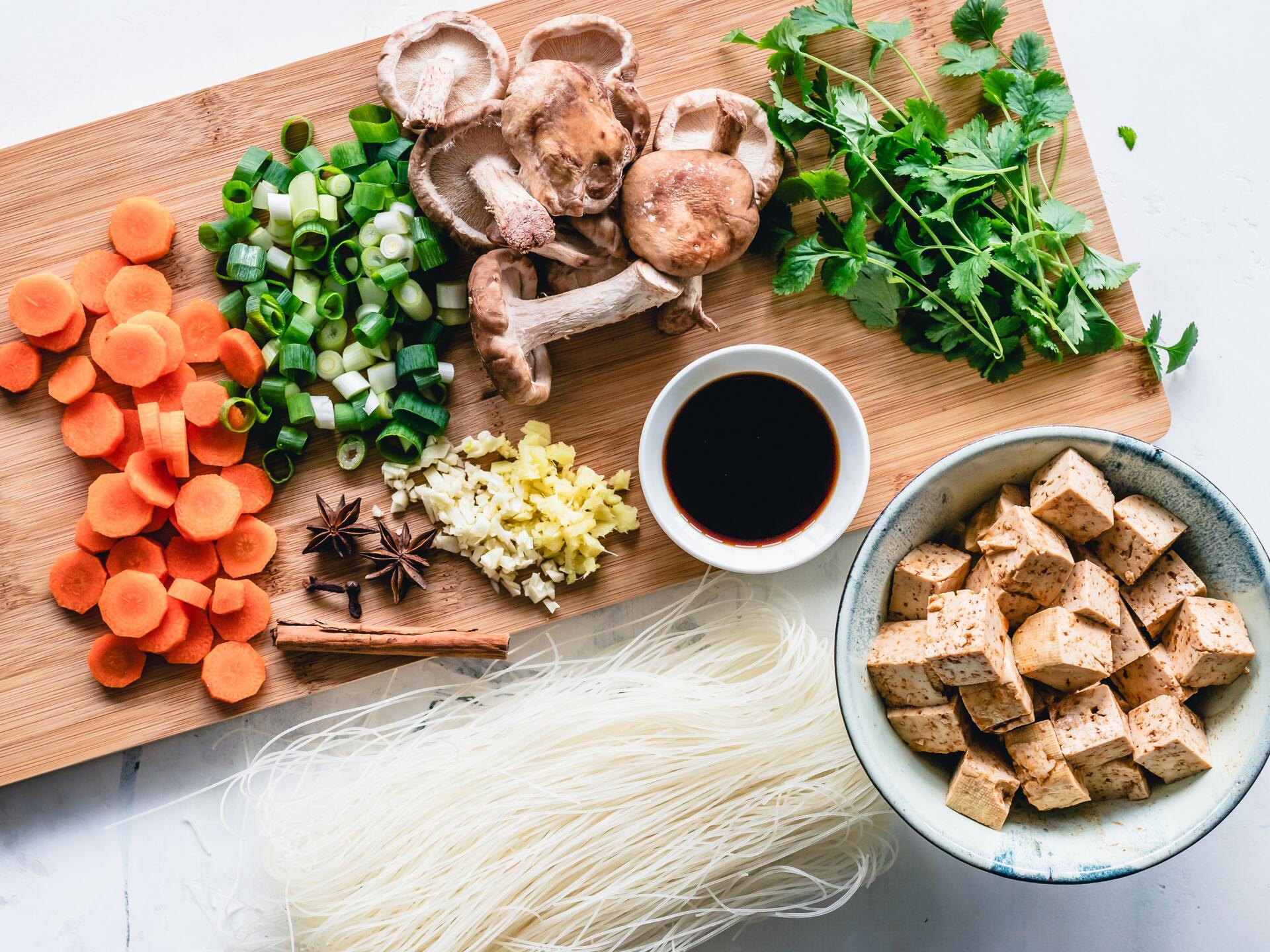 various foods and spices on a chopping board with mushrooms, rice noodles, and tofu in a bowl