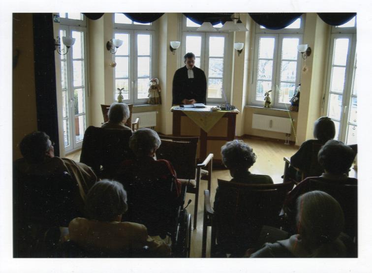 Dieses Bild zeigt den Geistlichen Bert Missal im Talar mit zum Gottesdienst versammelten Bewohnern eines Altersheimes.