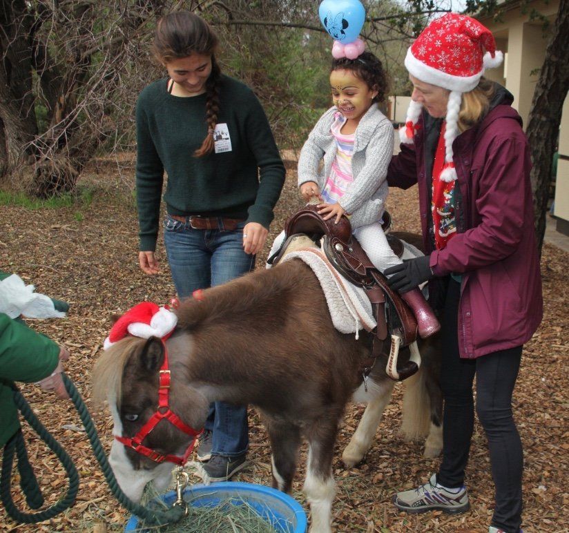 A smiling toddler riding a pony with the help of two volunteers.