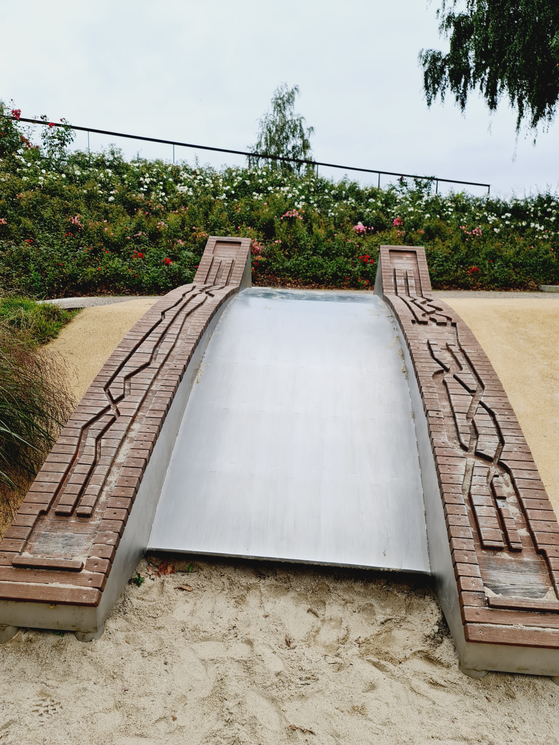 Lädt diese Rutsche nicht zum Mitmachen ein? Auch die Murmelbahn an den Seiten ist toll. Entdeckt im Bürgerpark Oelsnitz im Erzgebirge Photo by: Viola Einert-Krug breite Rutsche aus Metall mit Holzmurmelbahn an den Seite- liegt im Sand auf einem Hügel - oberhalb Blumen und Bäume