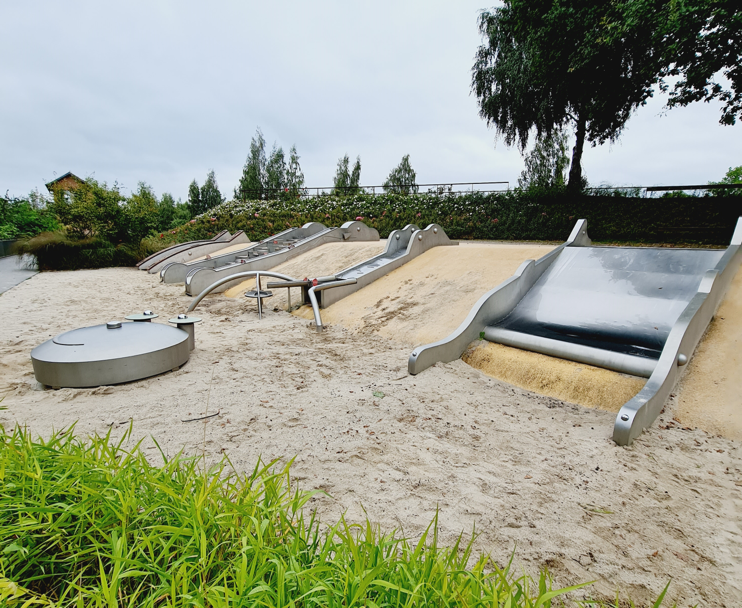 Rutschen, matschen, Spaß haben mit Wasser und Sand - eine Spieloase m Bürgerpark Oelsnitz Erzgebirge Photo by: Viola einert-Krug Wasserspielplatz auf einem Hügel. Spielelemente aus Metall zum Rutschen und Matschen im Wasser, Hintergrund Blumen und Bäume im Bürgerpark Oelsnitz Erzgebirge
