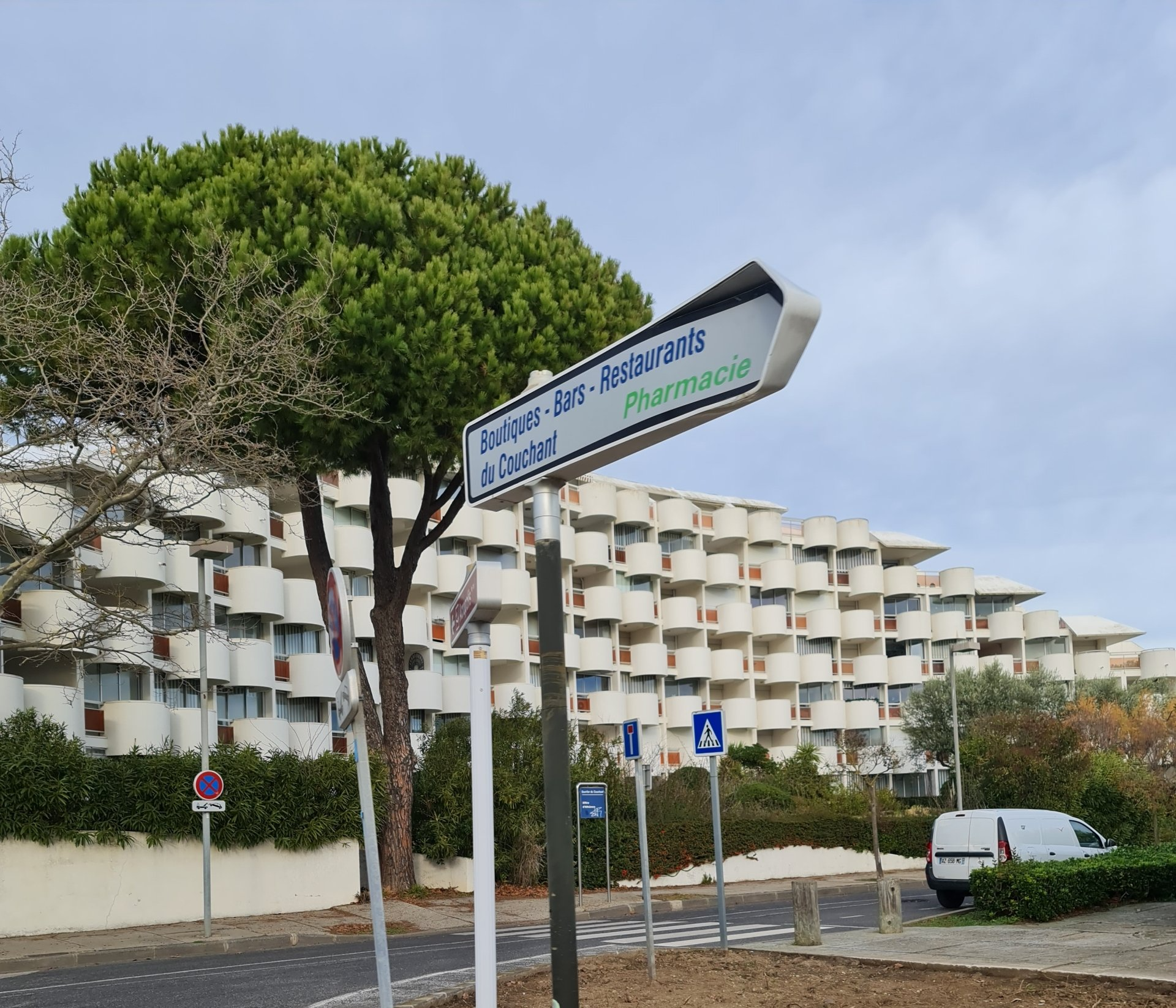Bäume vor einem im Halbkreis gebauten großen weißen Haus mit ausladend runden Balkons davor eine Straße mit Verkehrsschildern