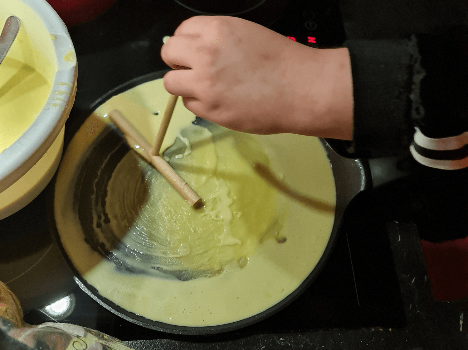 man sieht eine Hand die gerase Crepes in eine Pfanne zubereitet mit Holzsstäbchen