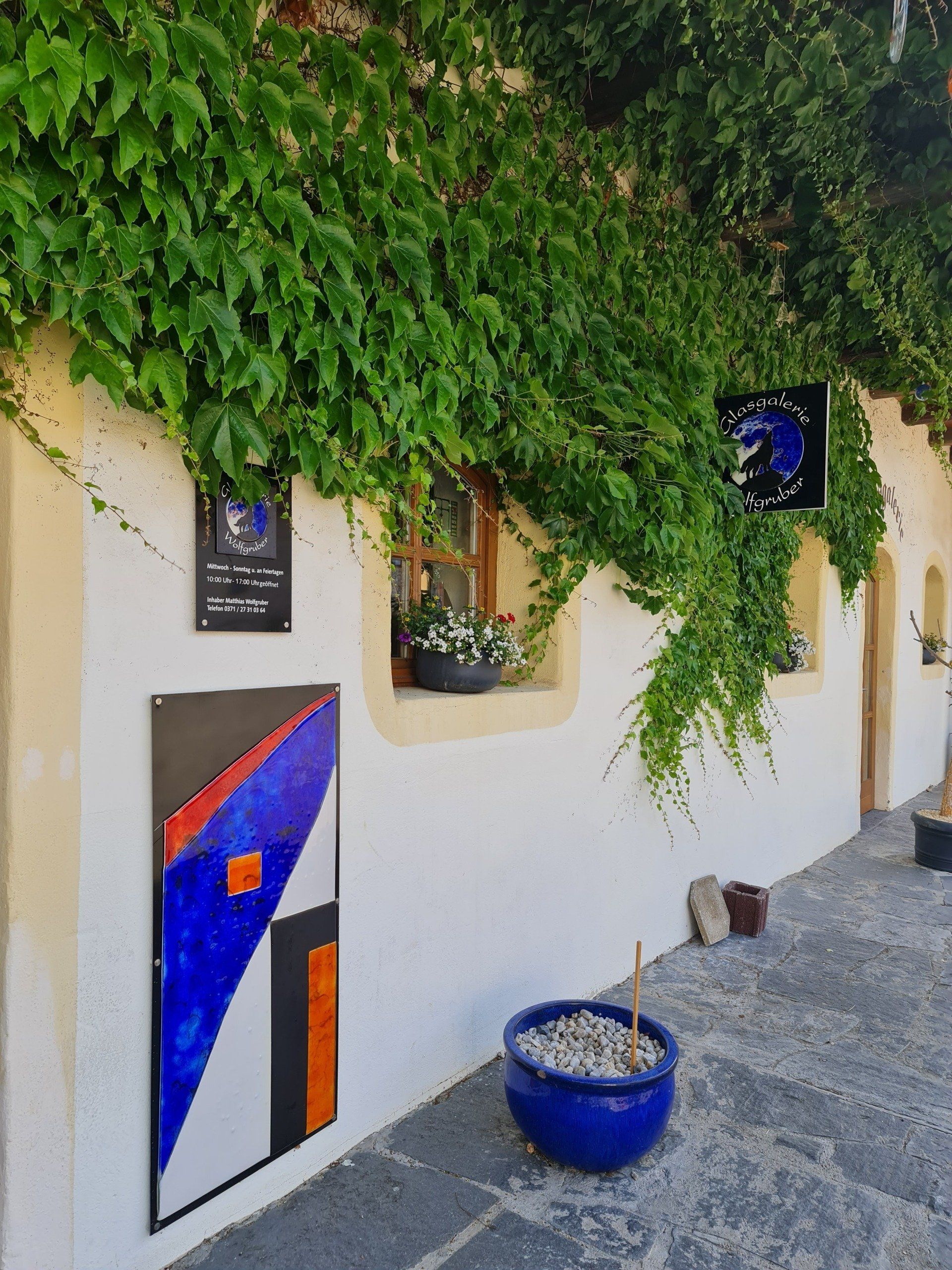 Eingang Glasgalerie mit Tafel aus Glas in blau weiß an der Wand in einem mit Wein bewachsenen Haus
