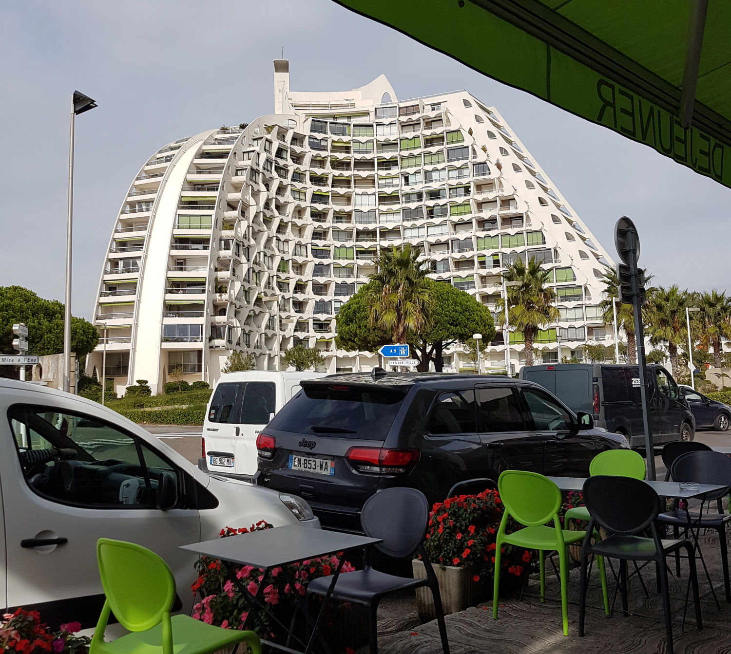 großes weißes Gebäude in einer pyramidenform gebaut mit vielen Fenstern und Balkonen, davor Sträucher und Autos